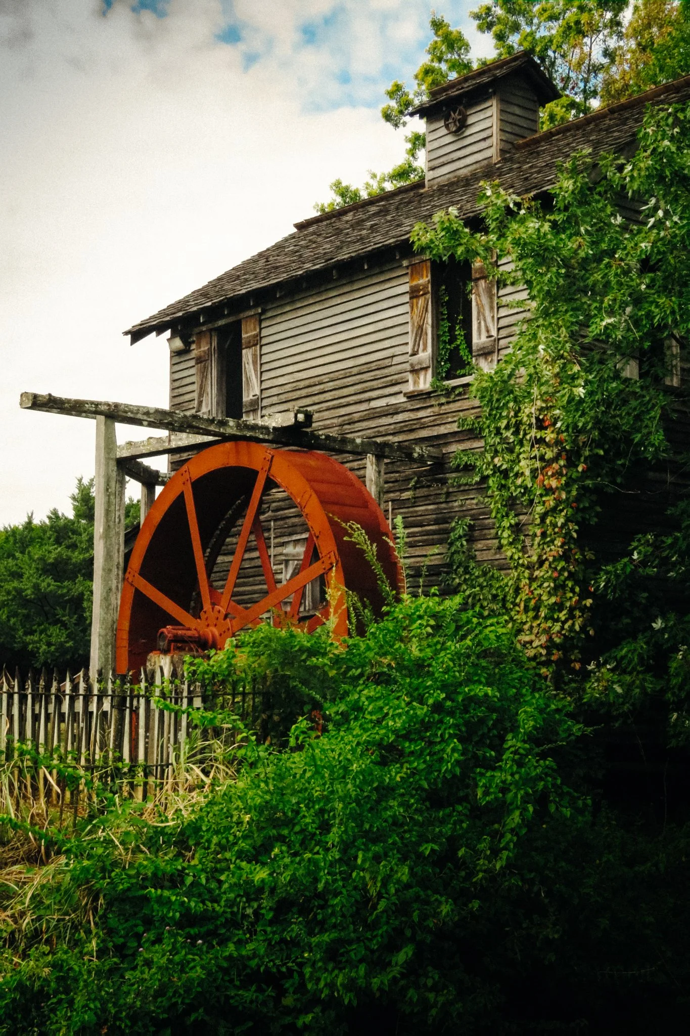 An old wooden barn with weathered shutters and a small vent on the roof. A large orange water wheel is in the foreground, partially obscured by lush green bushes and trees surrounding the barn.