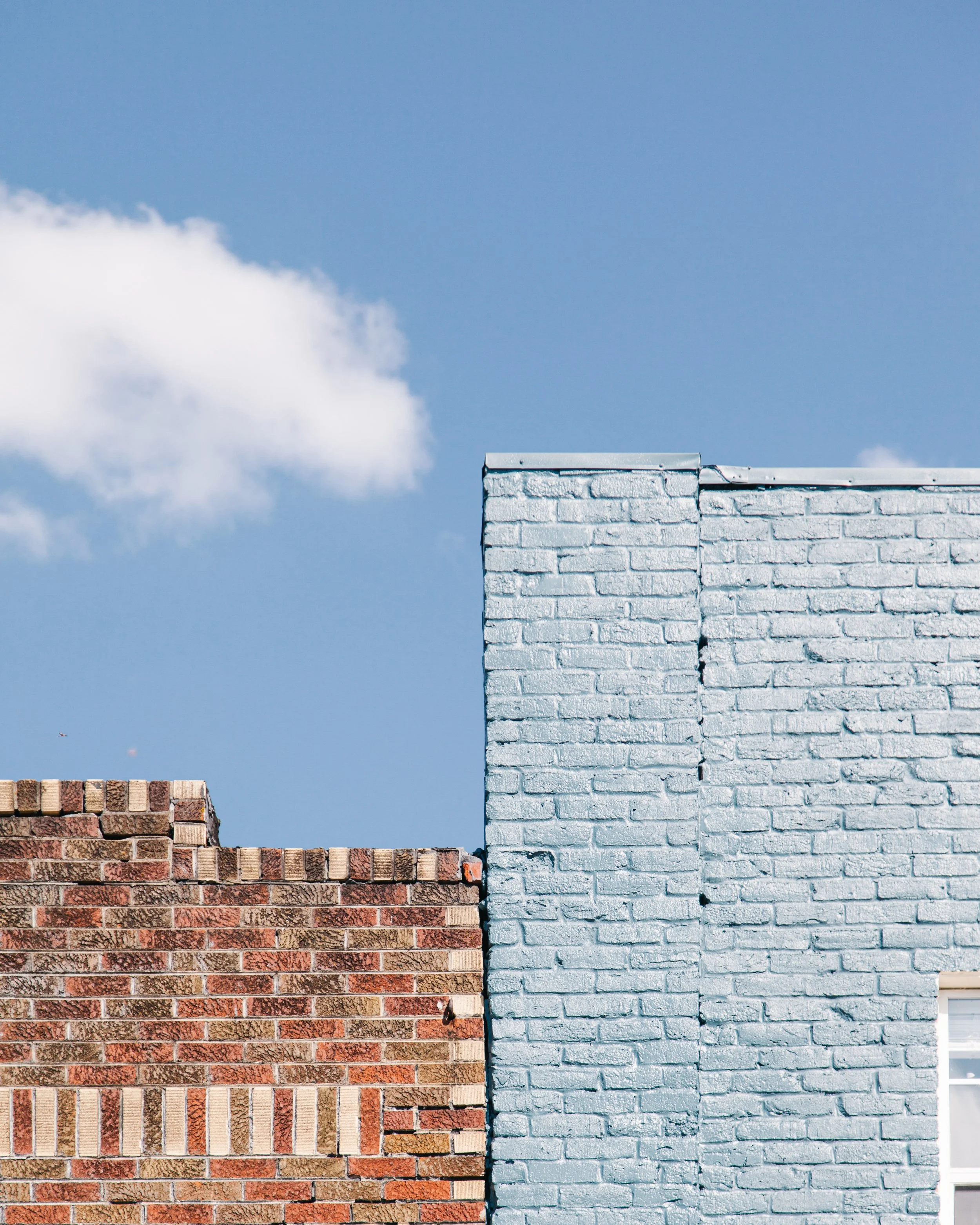 A wall of bricks with three different colors: the bottom bricks are reddish-brown, the middle bricks are light gray, and the top section is painted white. The a blue sky with some clouds is in the background.