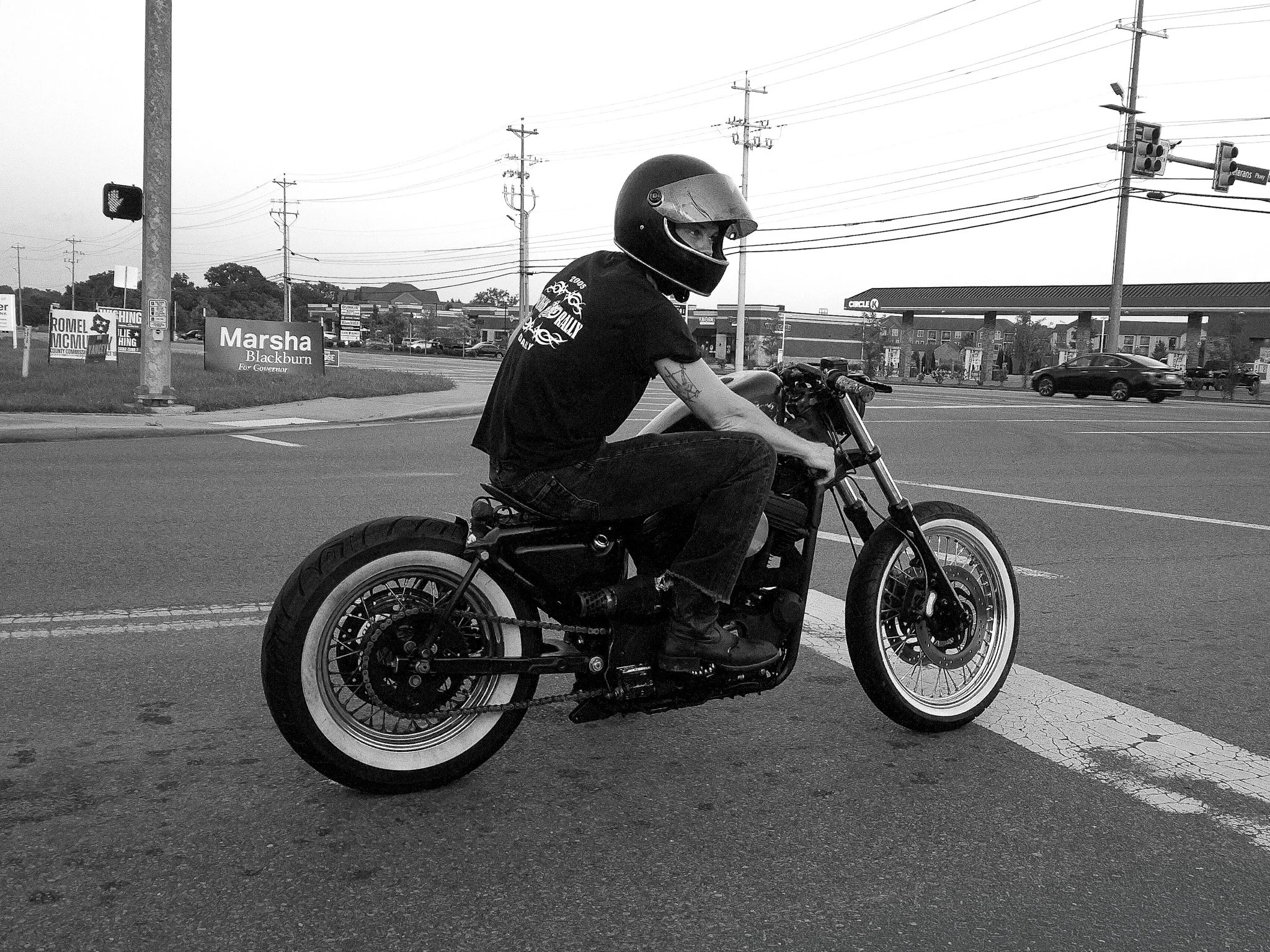 A person riding a motorcycle on a city street, wearing a black helmet and black clothing, with power lines, traffic lights, and commercial buildings in the background.