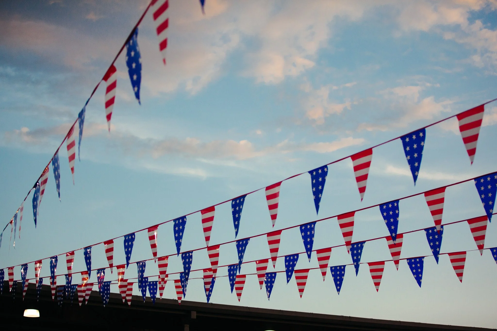 String of American flag-themed pennant banners hanging against a sky with scattered clouds.