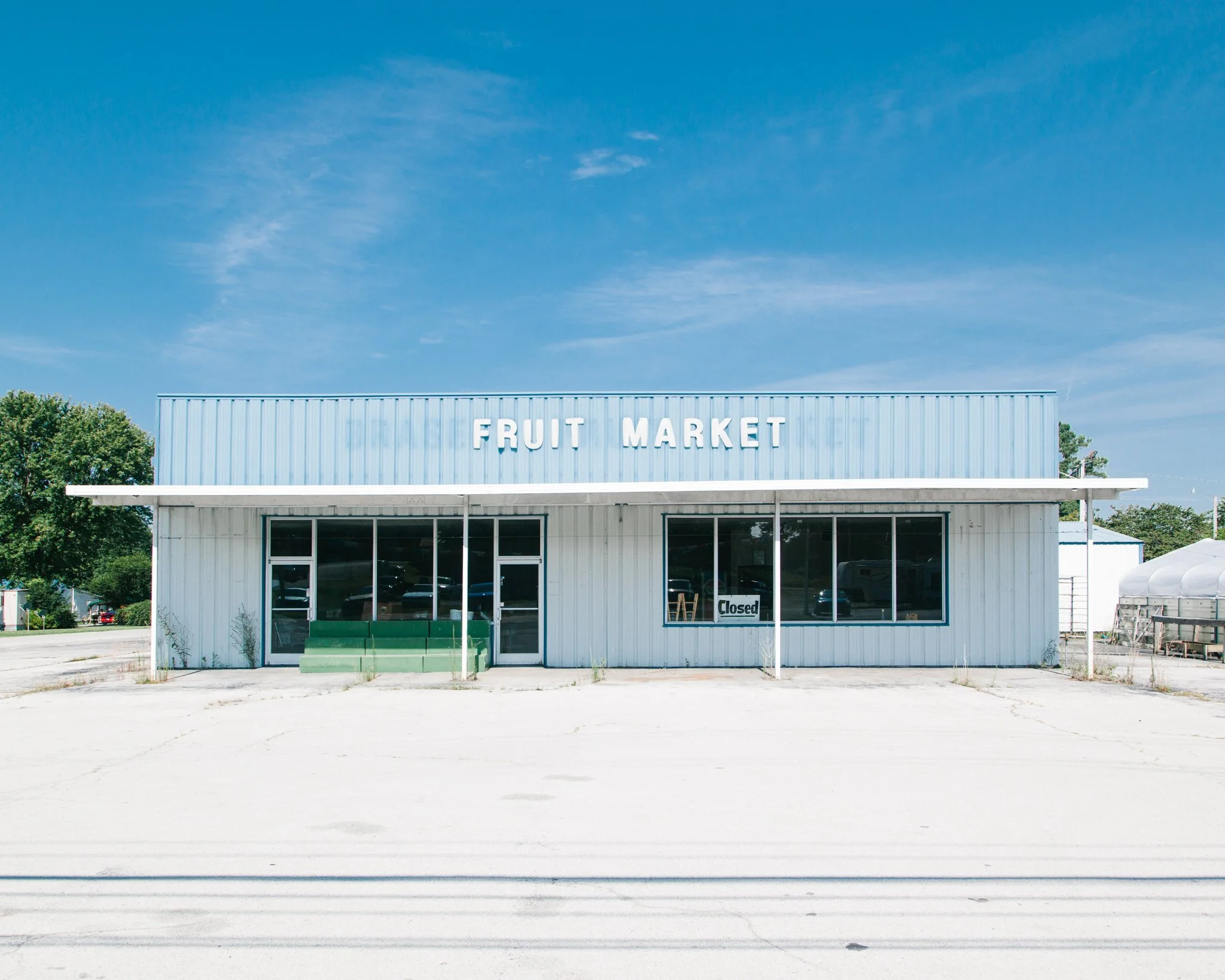 A white building with a blue roof and signage reading 'FRUIT MARKET' in white letters, situated under a bright blue sky with some clouds, with trees on either side and a cracked concrete area in front.