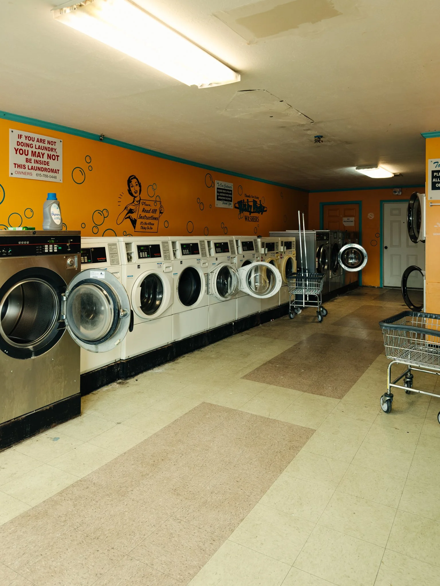 A laundromat with washing machines against a yellow wall with instructional posters and a shopping cart in the foreground.