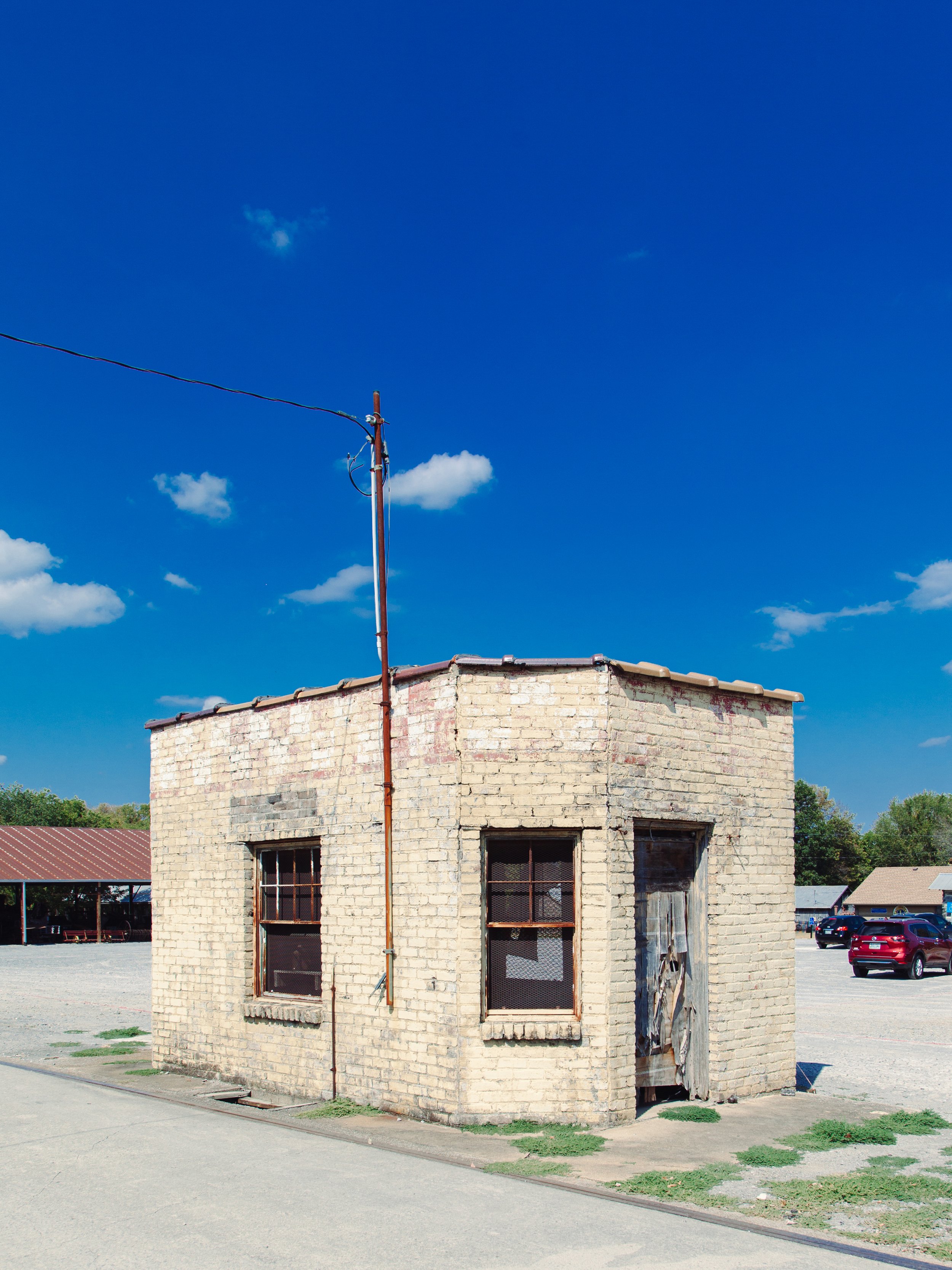 A small, old brick building with four windows, two of which are barred, and a damaged wooden door. The building is situated in a parking lot surrounded by cars and trees, under a bright blue sky with a few white clouds.