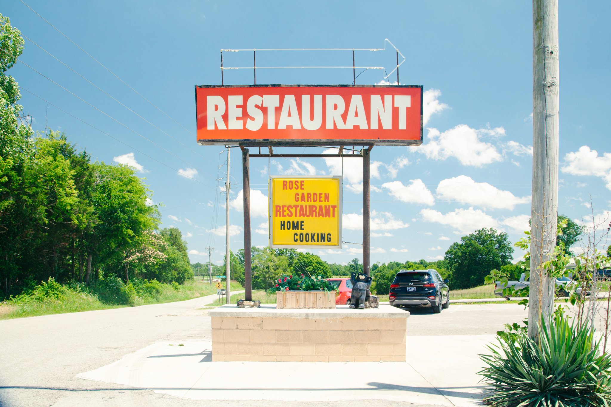 Outdoor parking lot with a large red sign that says 'RESTAURANT' and a smaller yellow sign promoting 'ROSE GARDEN RESTAURANT HOME COOKING'. Several parked cars are visible, along with trees and blue sky with white clouds.