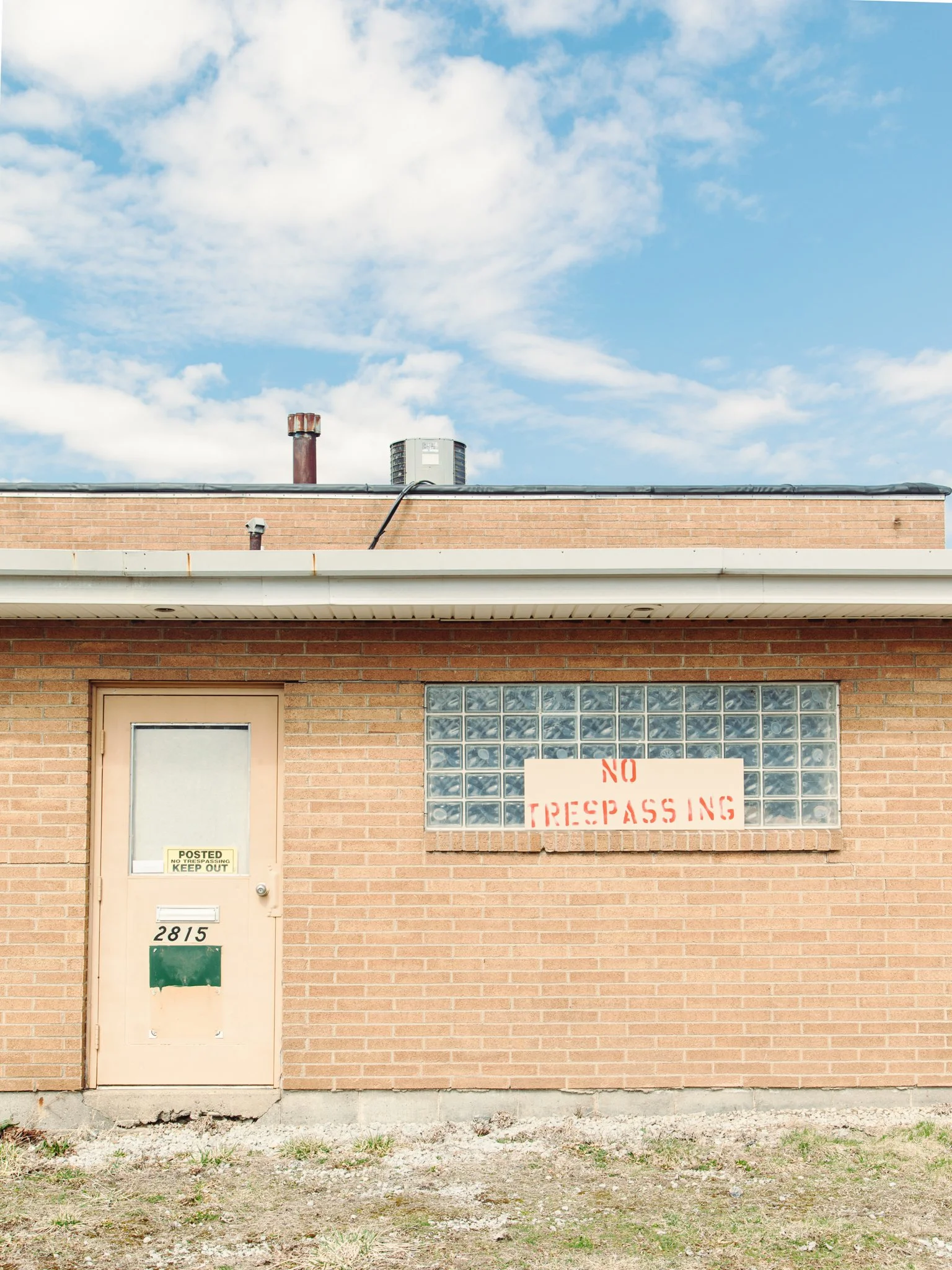 The image shows the exterior of a brick building with a door, a glass block window, and signs that read 'No Trespassing' and 'Posted No Trespassing Keep Out.' The ground in front is uneven with dirt and small patches of grass, and the sky above is pa