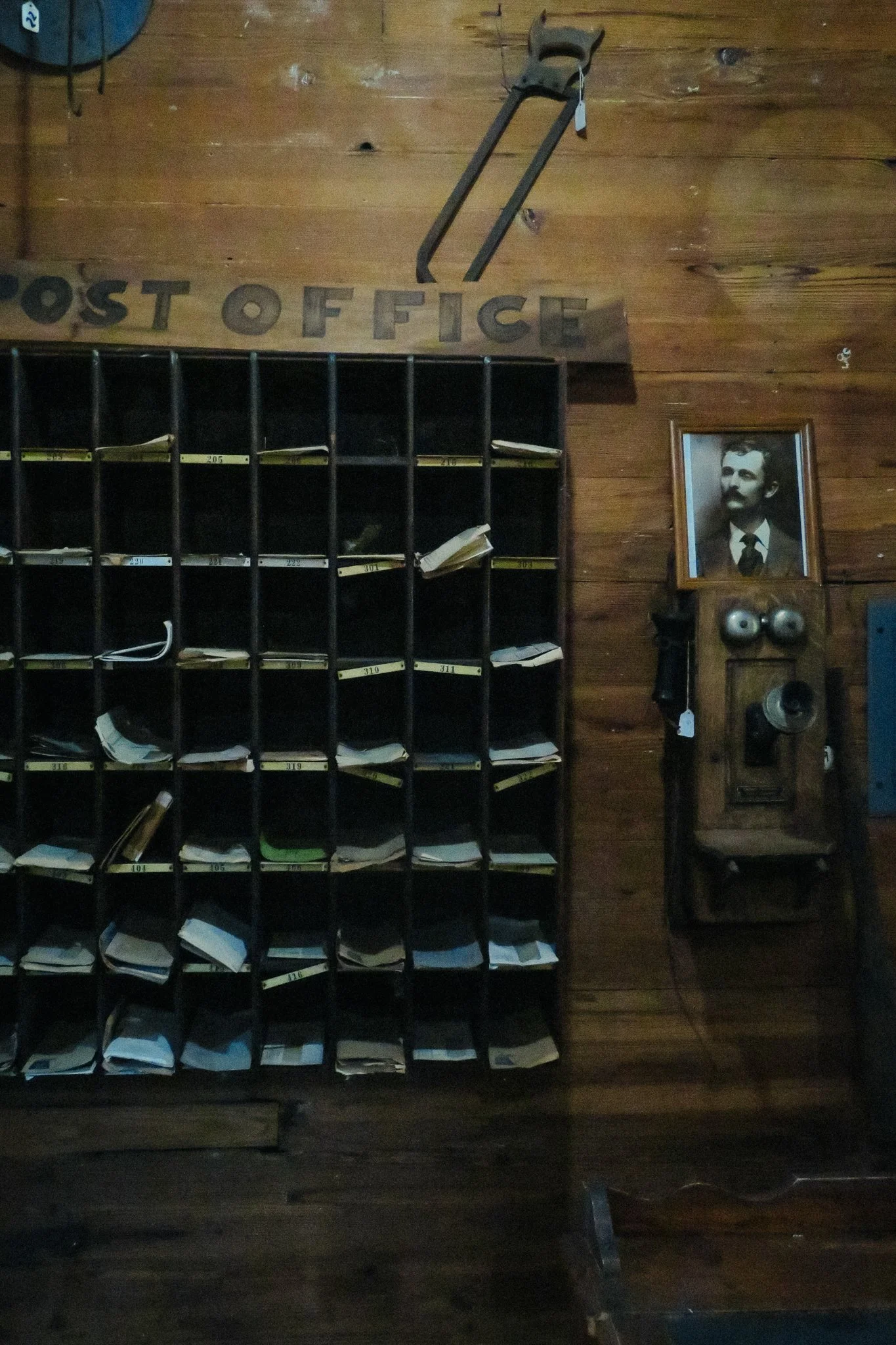 A wooden post office wall with a vintage clock, a saw hanging, a framed black-and-white photograph of a man with a mustache, a set of mailboxes with papers and envelopes inside, and a wooden cabinet with a vintage telephone and clock.