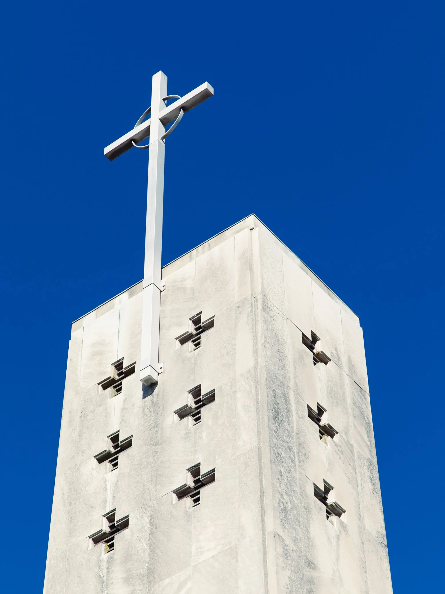 A white modern building with small square windows against a clear blue sky, with a tall white cross structure in the foreground.