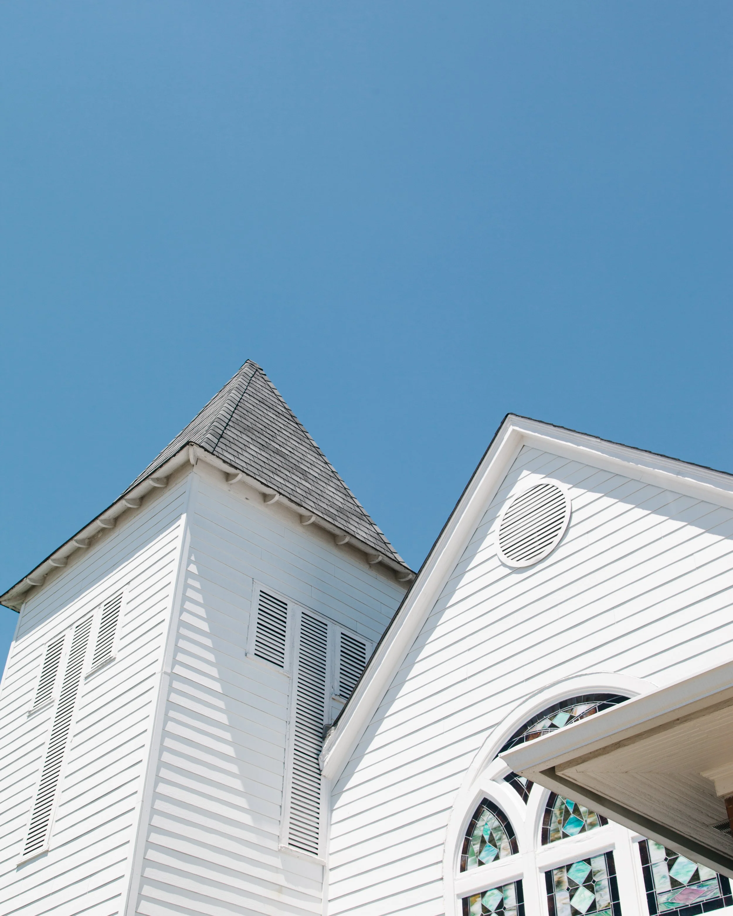 Close-up of the upper part of a white church building with pointed steeples, decorative windows with stained glass, and siding, under a clear blue sky.