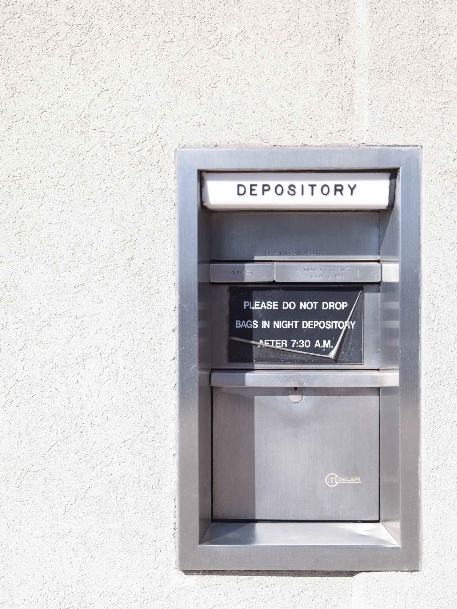 A metal deposit box mounted on a beige stucco wall, with a sign indicating it is a deposit box for dropping off bags after 7:30 A.M.