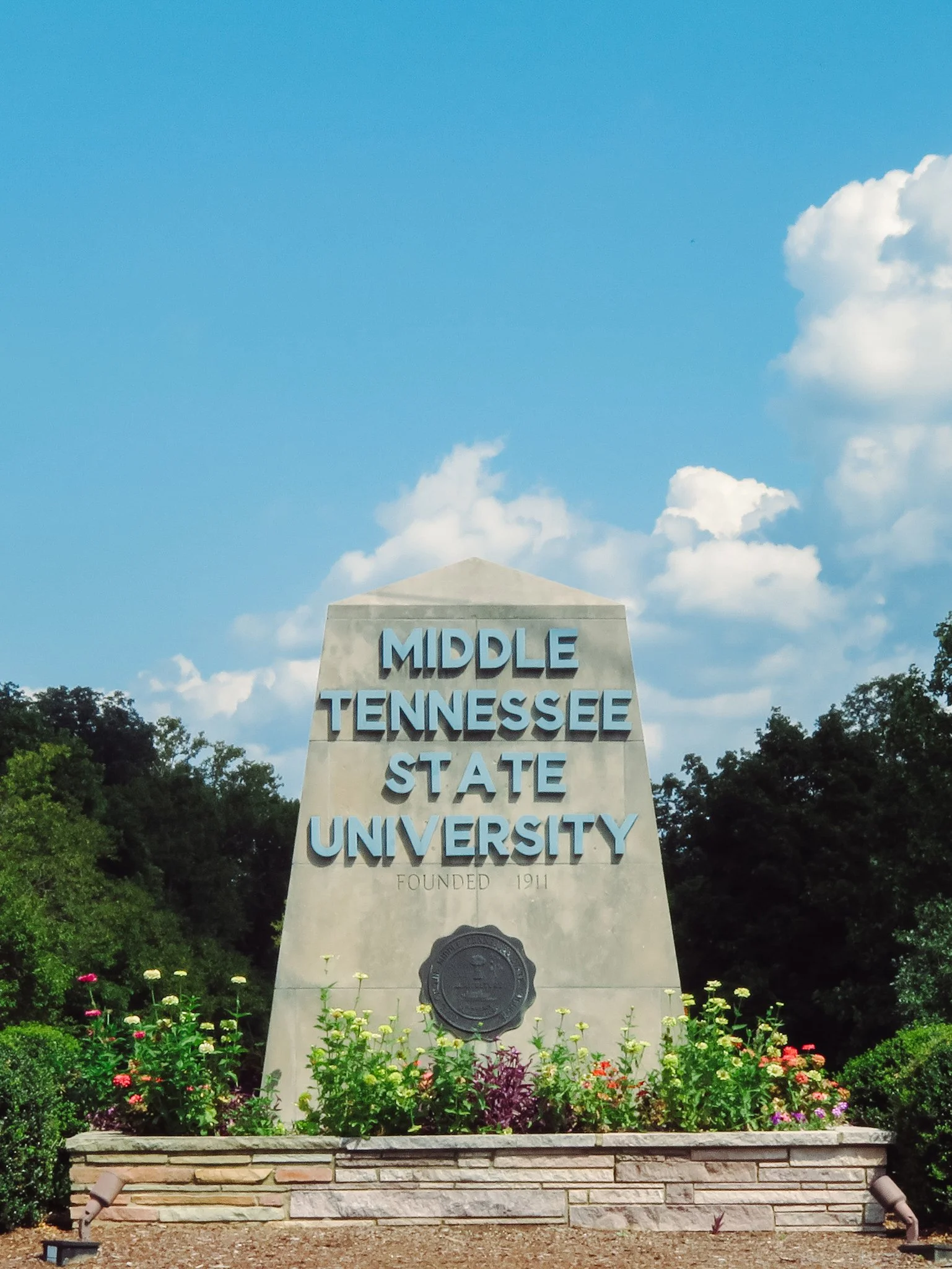 Stone monument with the inscription 'Middle Tennessee State University, founded 1911' surrounded by flower bed and greenery under blue sky with clouds.