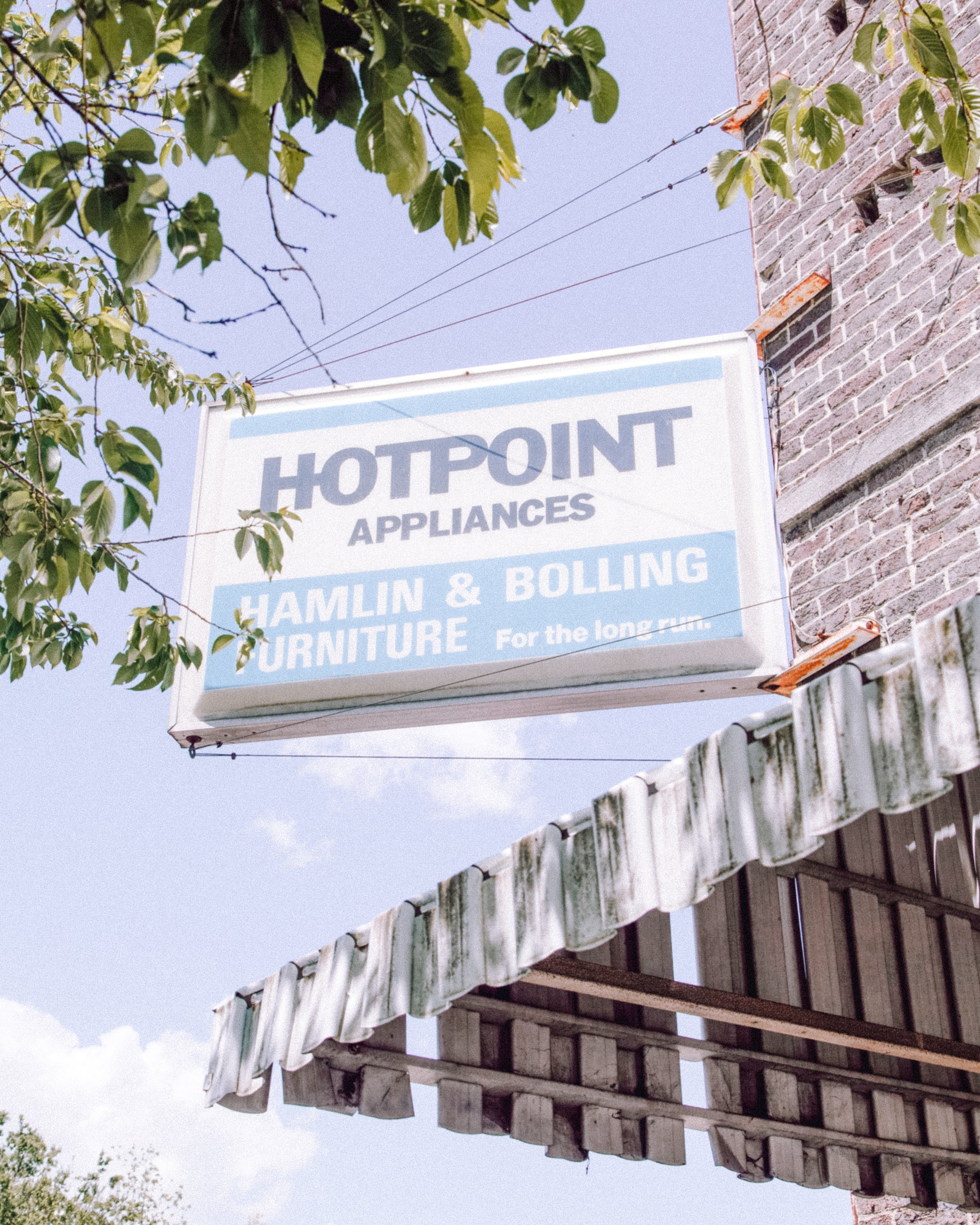 A sign for Hotpoint appliances hanging on the side of a brick building, with a tree branch with green leaves partially obscuring the sign. The sky is visible in the background.