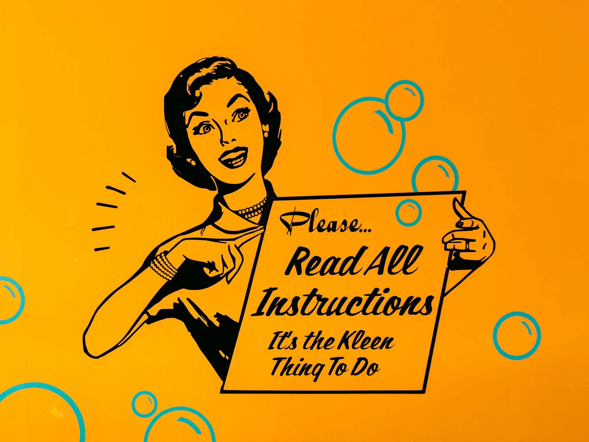 A pop art style illustration of a woman holding a sign that says 'Please Read All Instructions It the Kleen Thing To Do.' The woman has dark hair, is wearing a necklace and bracelet, and has an animated expression with one eye winking. The background