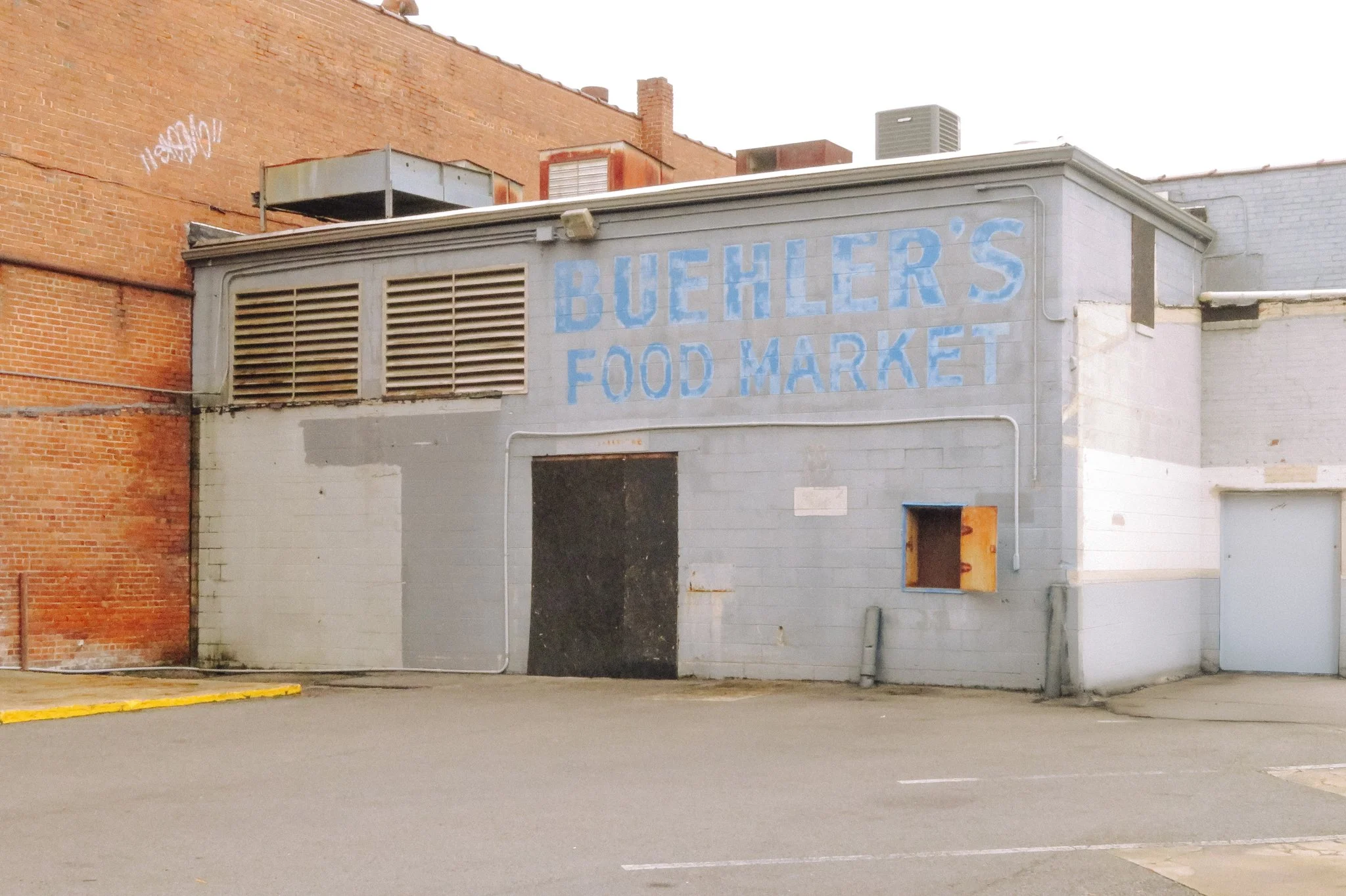 A building with gray painted exterior and faded blue lettering that reads 'BUEHLER'S FOOD MARKET.' The building is adjacent to a red brick wall with some graffiti on it. There are no people or vehicles visible.