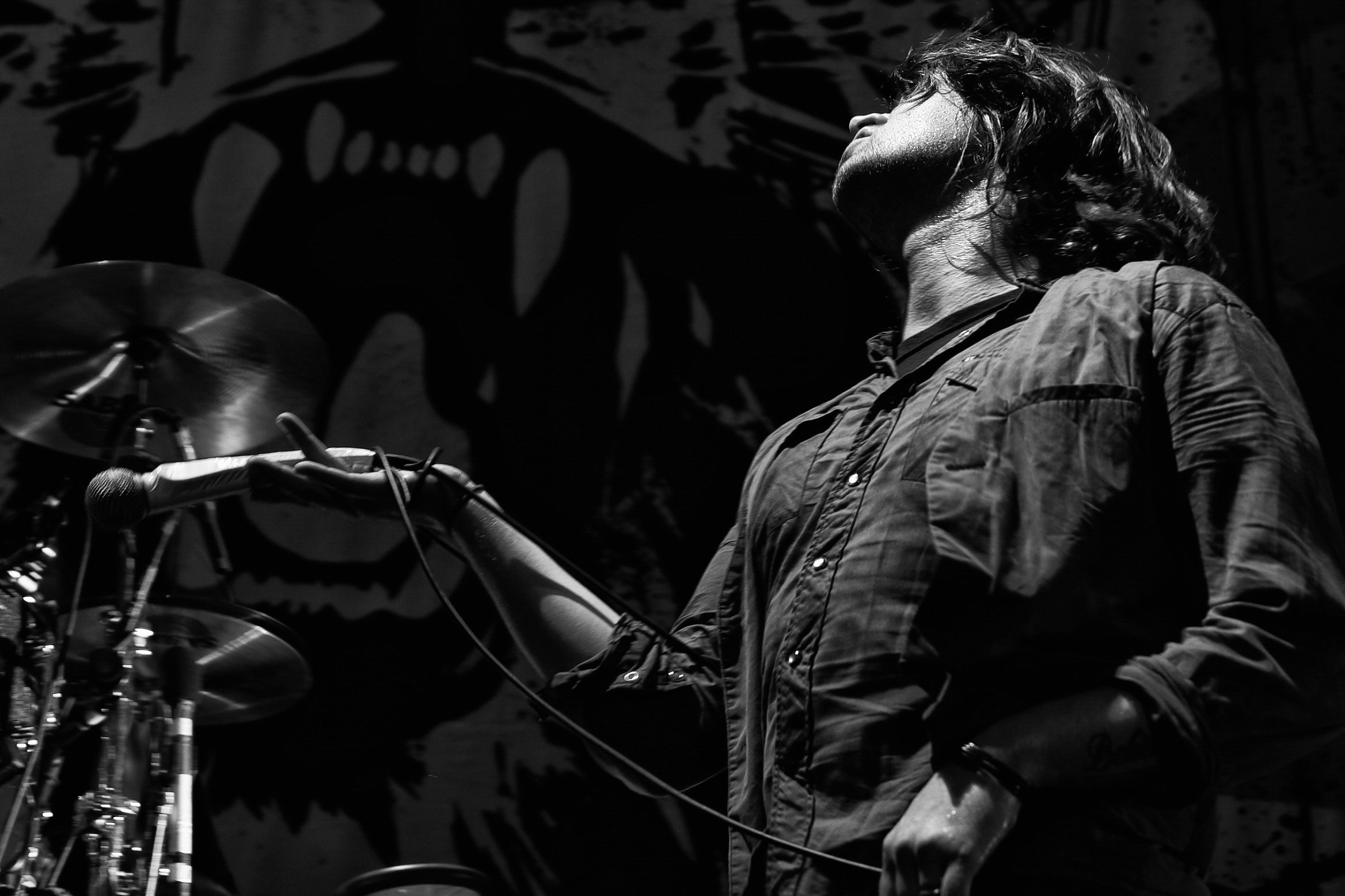 A person with styled curly hair wearing a button-up shirt, seen in profile, standing next to a motorcycle in a dimly lit environment with shadows and background patterns.