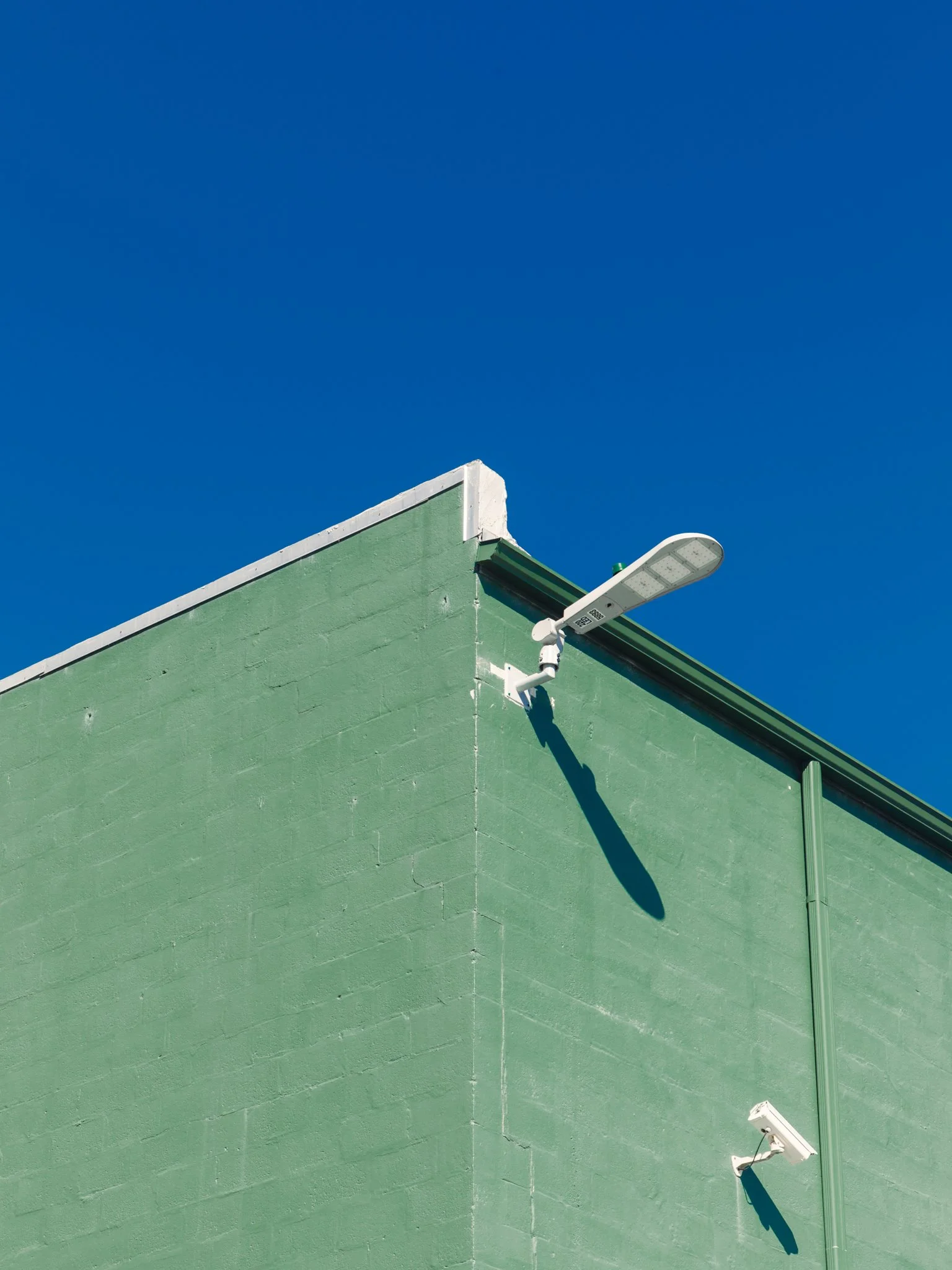 A corner of a green building with a dark green trim, with two white security cameras and an outdoor LED streetlight mounted on the wall, against a clear blue sky.