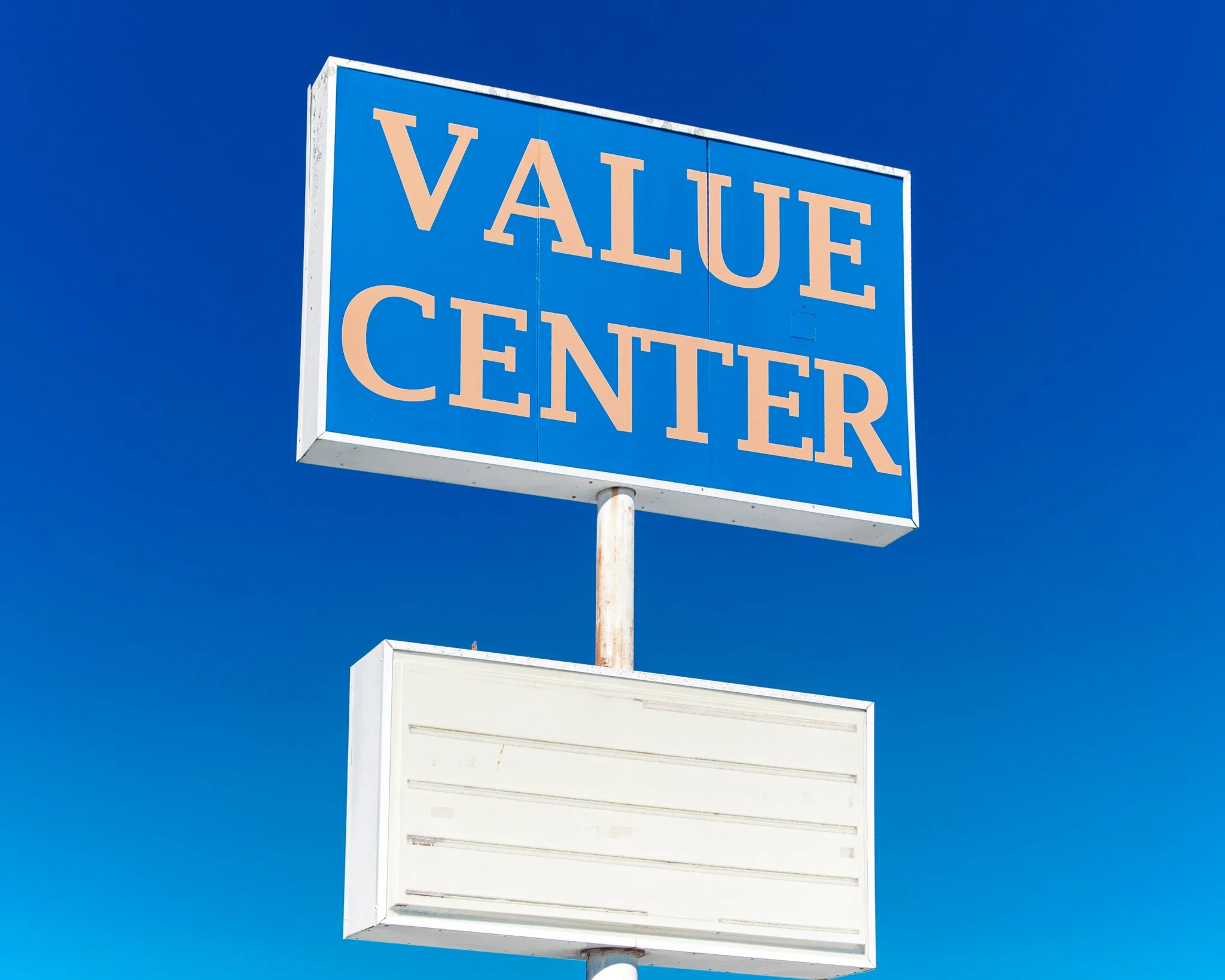 A blue rectangular sign with the words "VALUE CENTER" in large peach-colored letters against a clear blue sky. The sign appears to be on a tall pole, with the lower part of the sign blank and the upper part displaying the text.