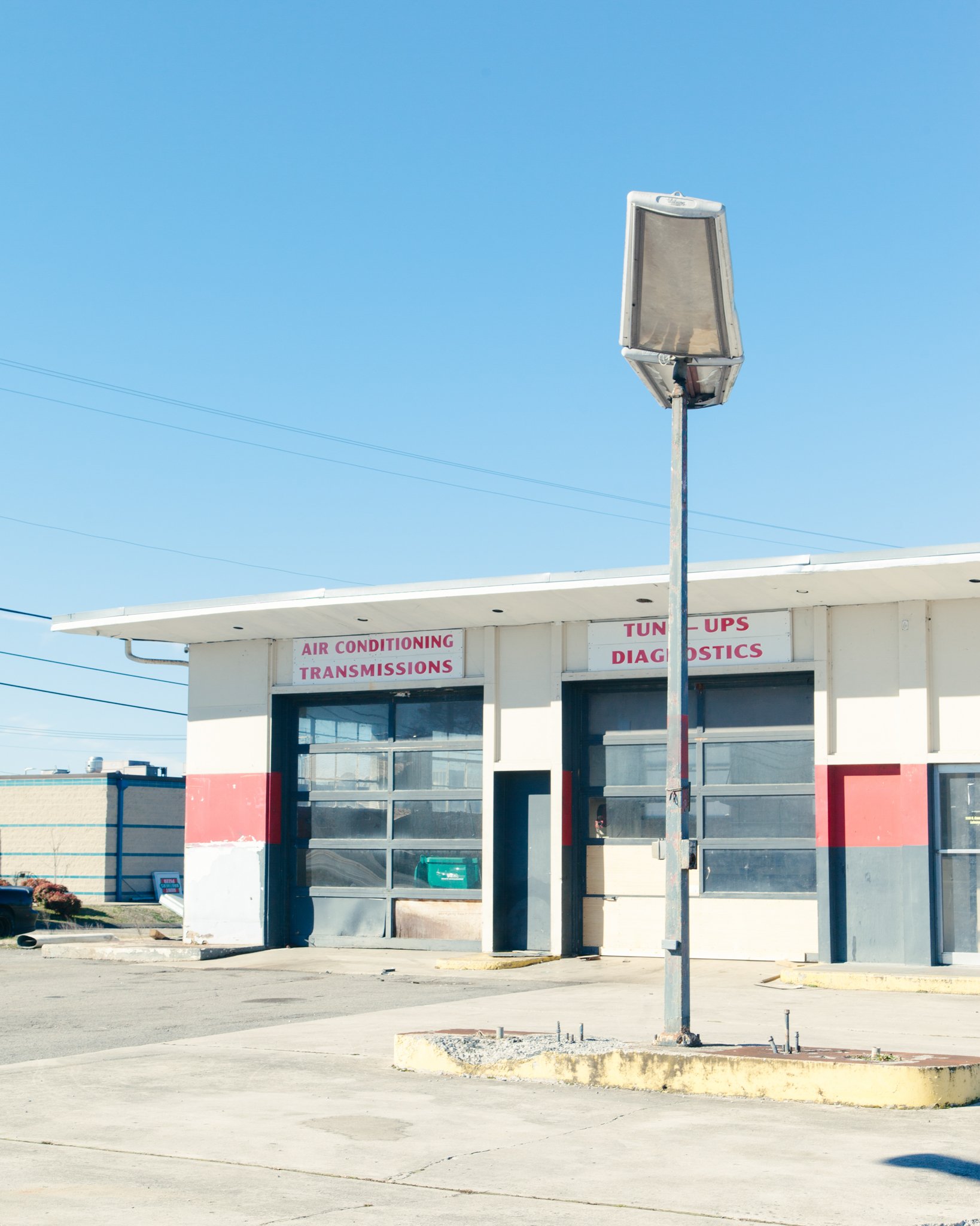 Empty building with signs for air conditioning, transmissions, tune-ups, and diagnostics, and a large streetlamp in front, under a clear blue sky.
