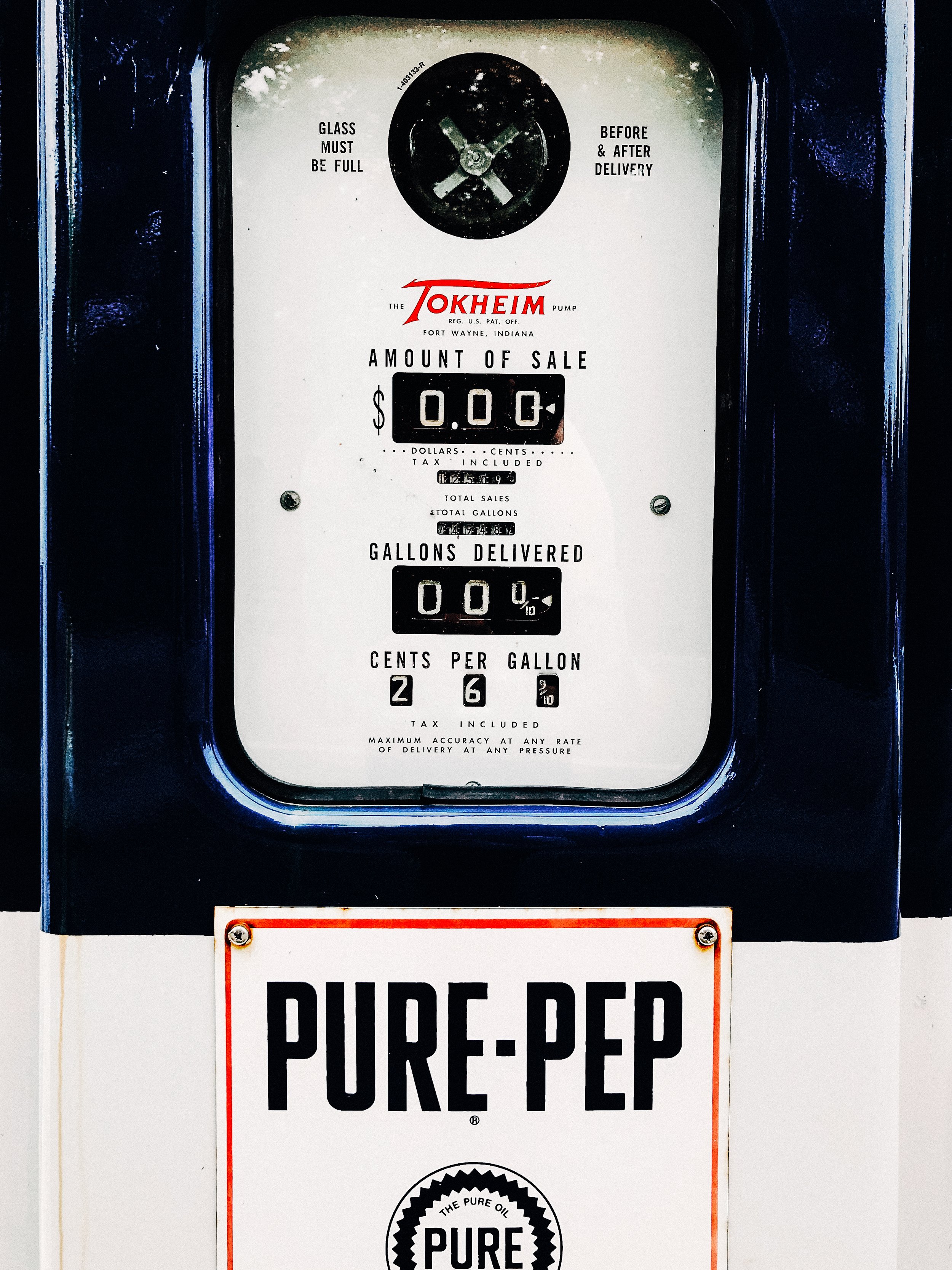 A vintage oil pump with a dial showing zero gallons delivered and the sign below reading 'PURE-Pep' in bold black letters on a white background, with a decorative seal stating 'The Pure Oil'.