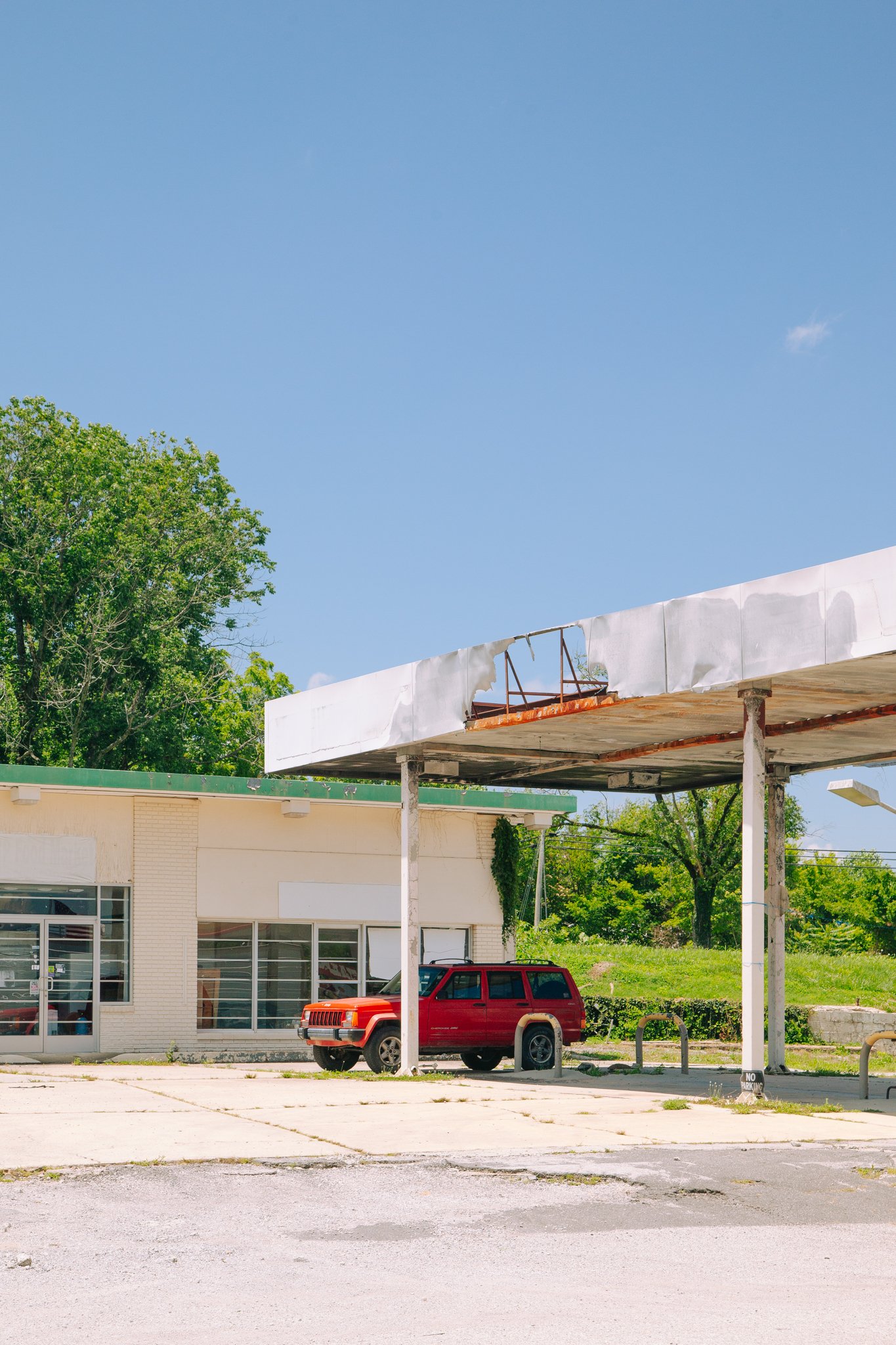 A red Jeep parked in front of a building with large windows, under a white overhang, with a green roof and tree in the background, and a clear blue sky overhead.
