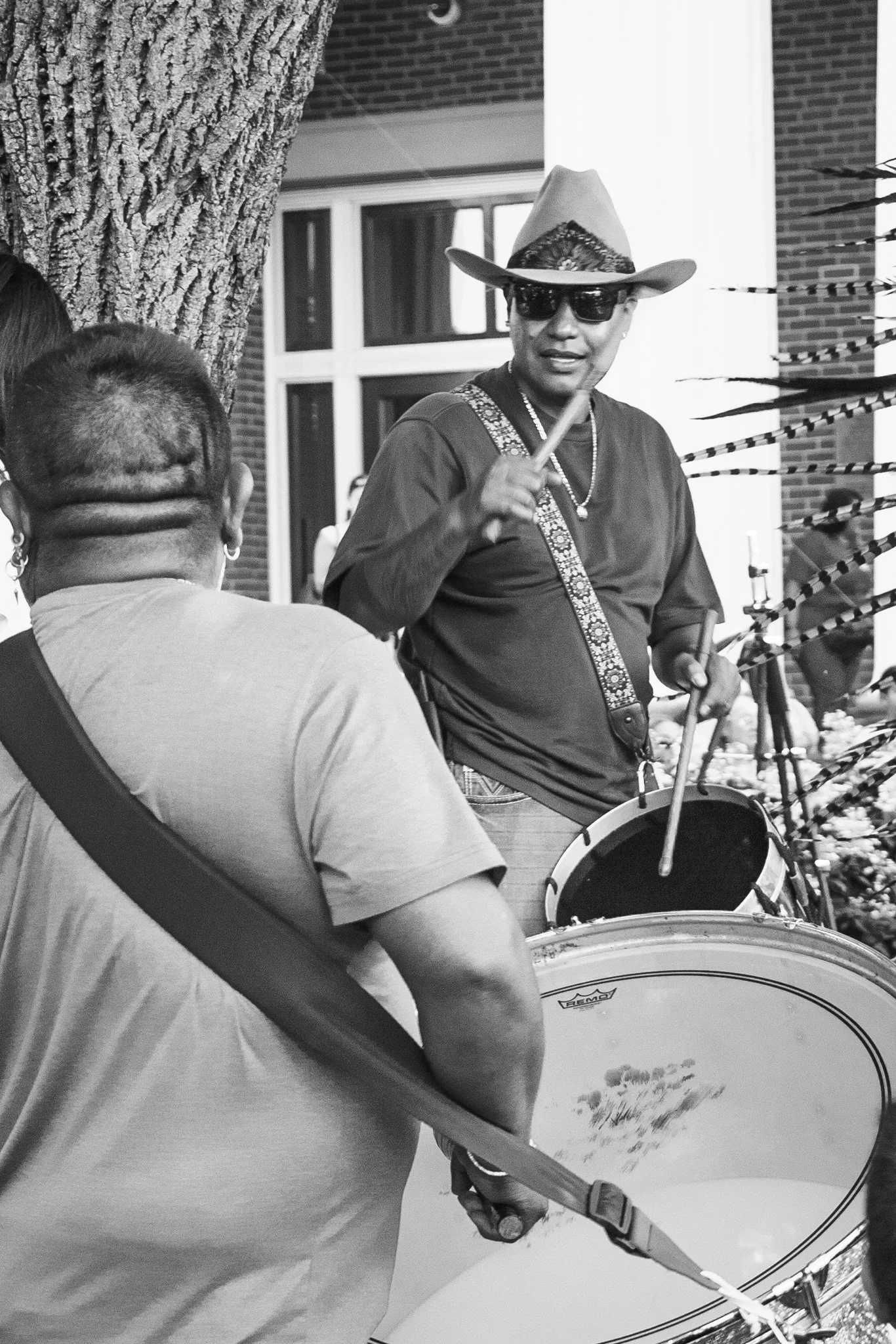 A man wearing sunglasses and a cowboy hat is playing drums outdoors near a brick building and a tree.