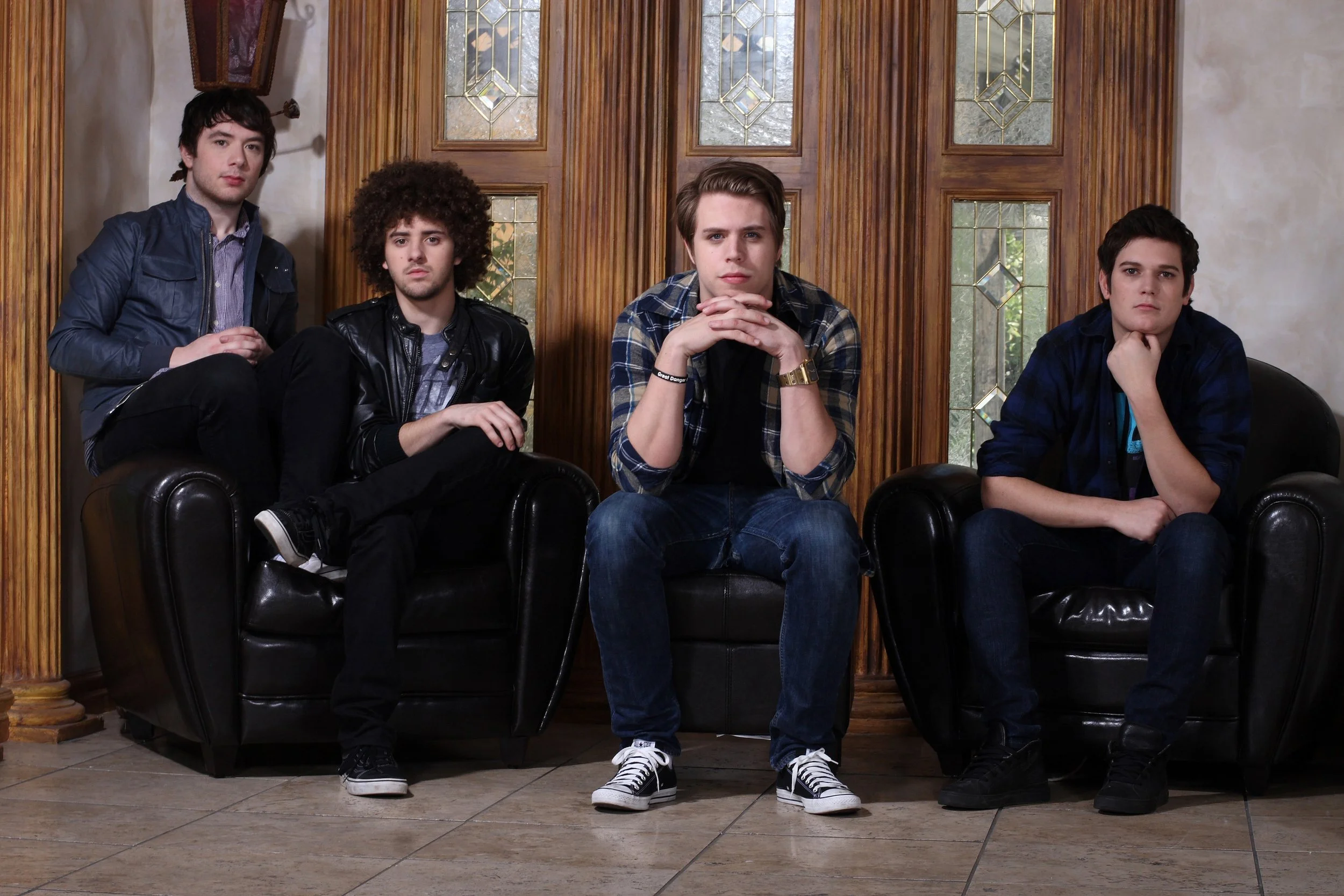 Four young men sitting on black leather chairs in a room with wood-paneled walls and stained glass windows. They appear serious and are looking directly at the camera.