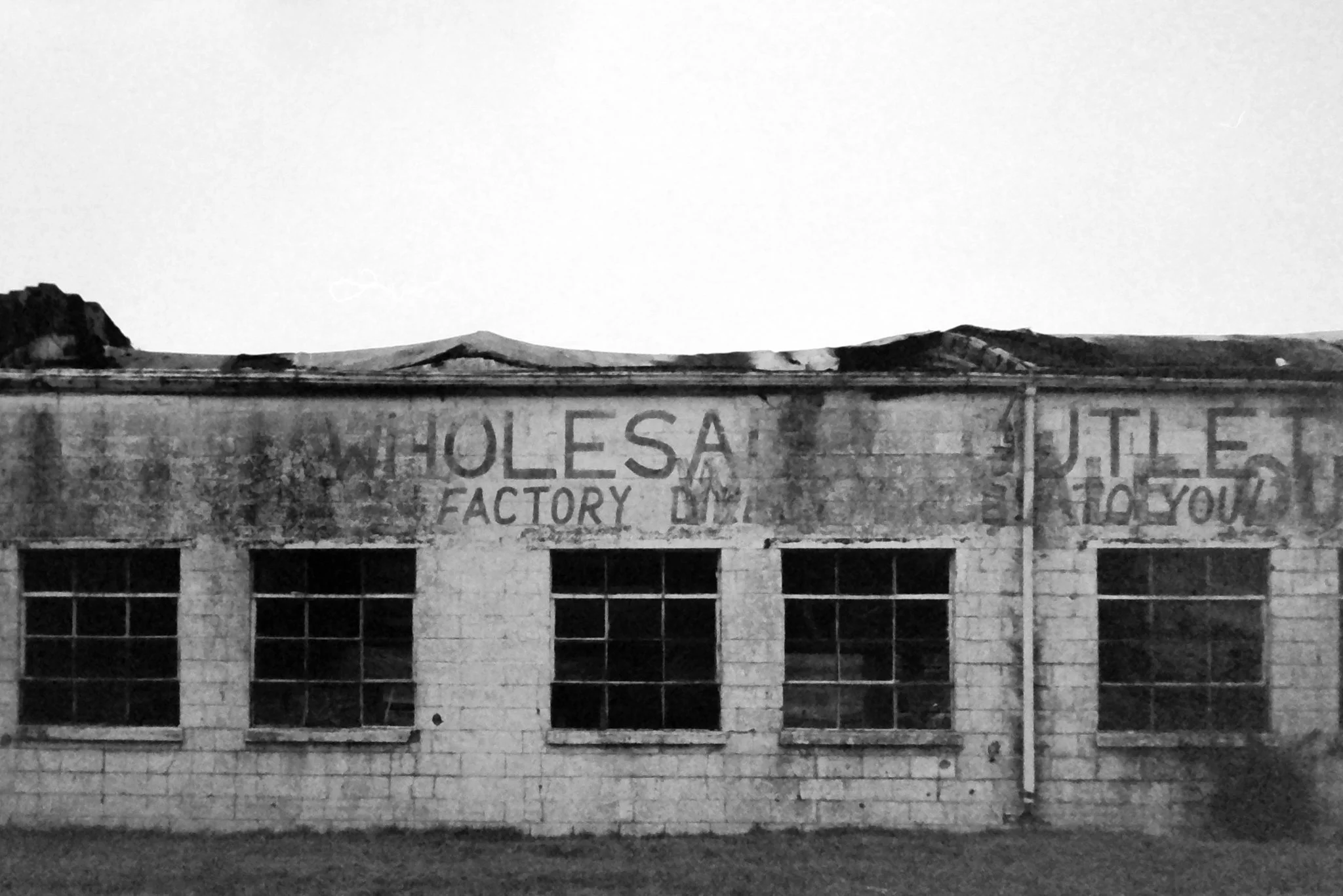 An old brick building with a faded painted sign reading 'H.O.L.E.S. Factory' on the top facade. The building has multiple windows and is set against a clear sky with distant hills.