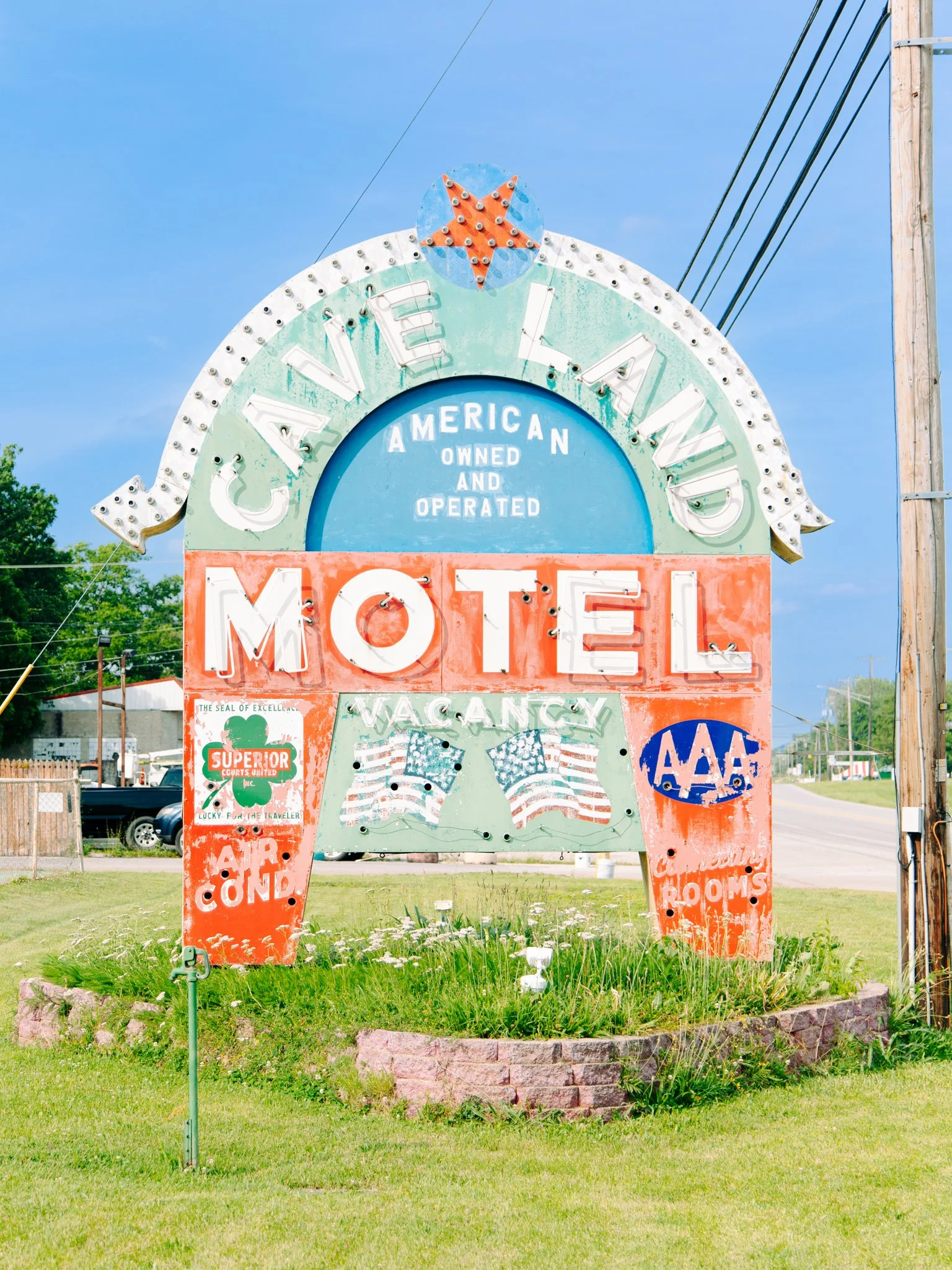 Vintage roadside sign reading 'Travel American Owned and Operated' with a large 'Motel' sign, decorated with patriotic American flags and advertisements, set in a grassy area near a residential or commercial street.