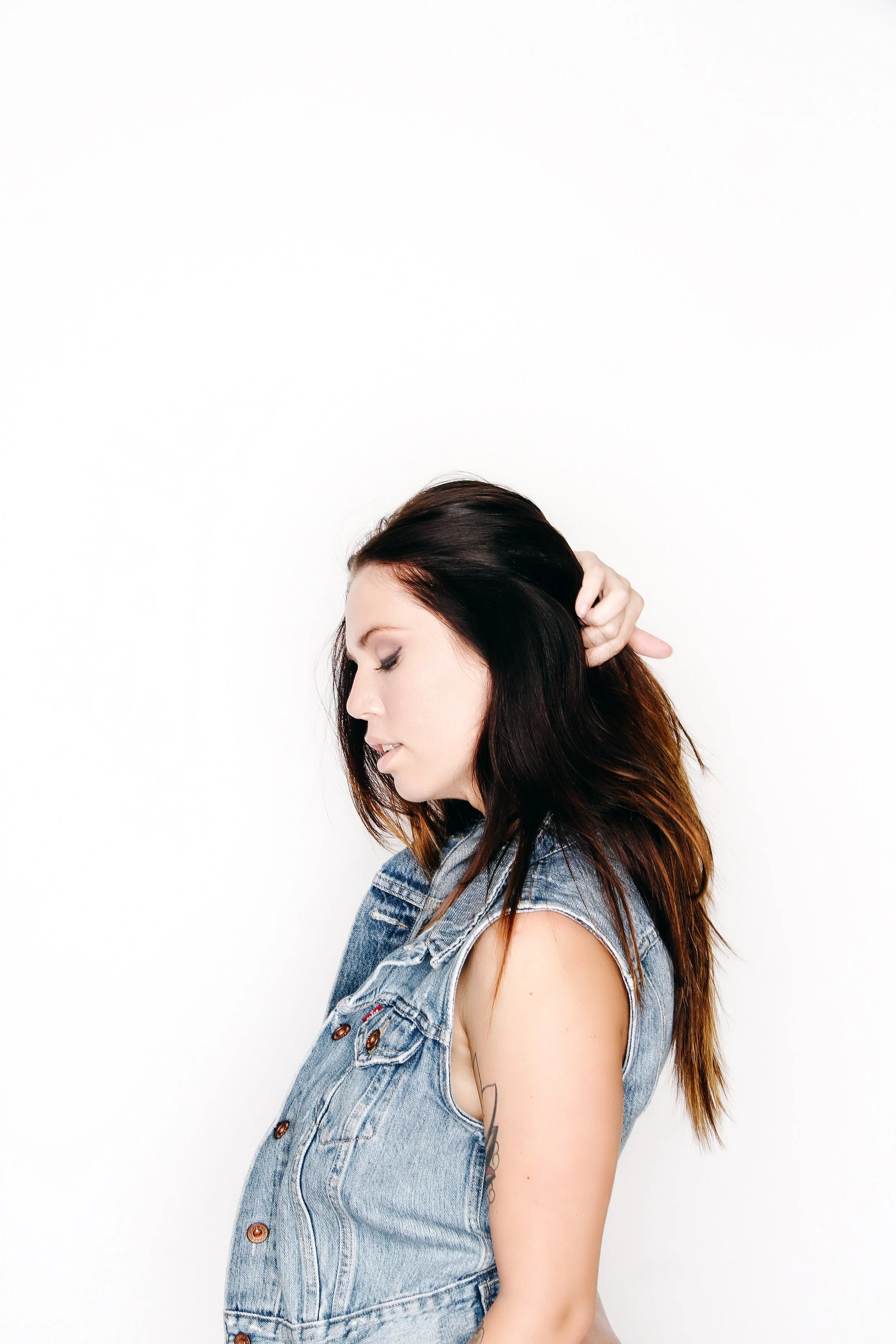 A woman with long brown and black hair wearing a ripped denim vest, eyes closed, head tilted downward, against a plain white background.