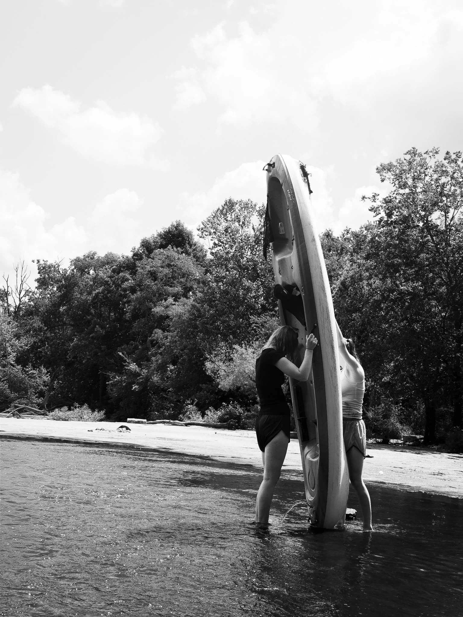 Two women stand in shallow water, helping a person secure a kayak to the kayak rack during daytime, with trees and clouds in the background.