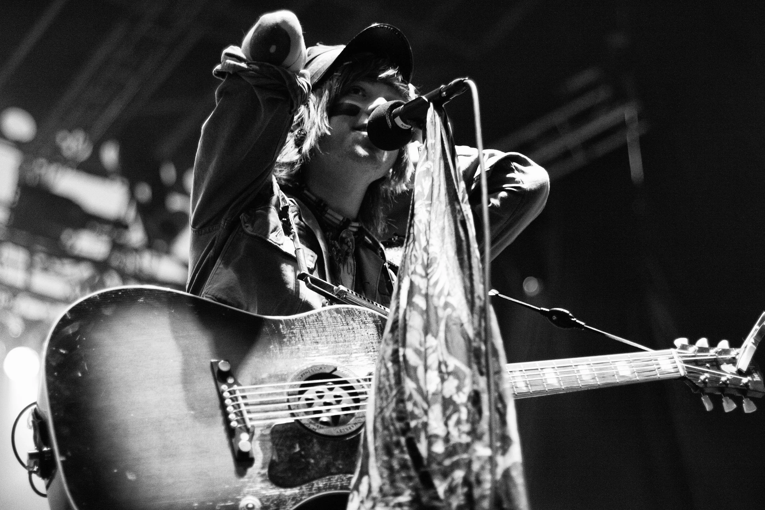 A person with long hair wearing a hat and face paint, performing on stage with a microphone, playing an acoustic guitar, with a patterned cloth on the microphone stand, in black and white.