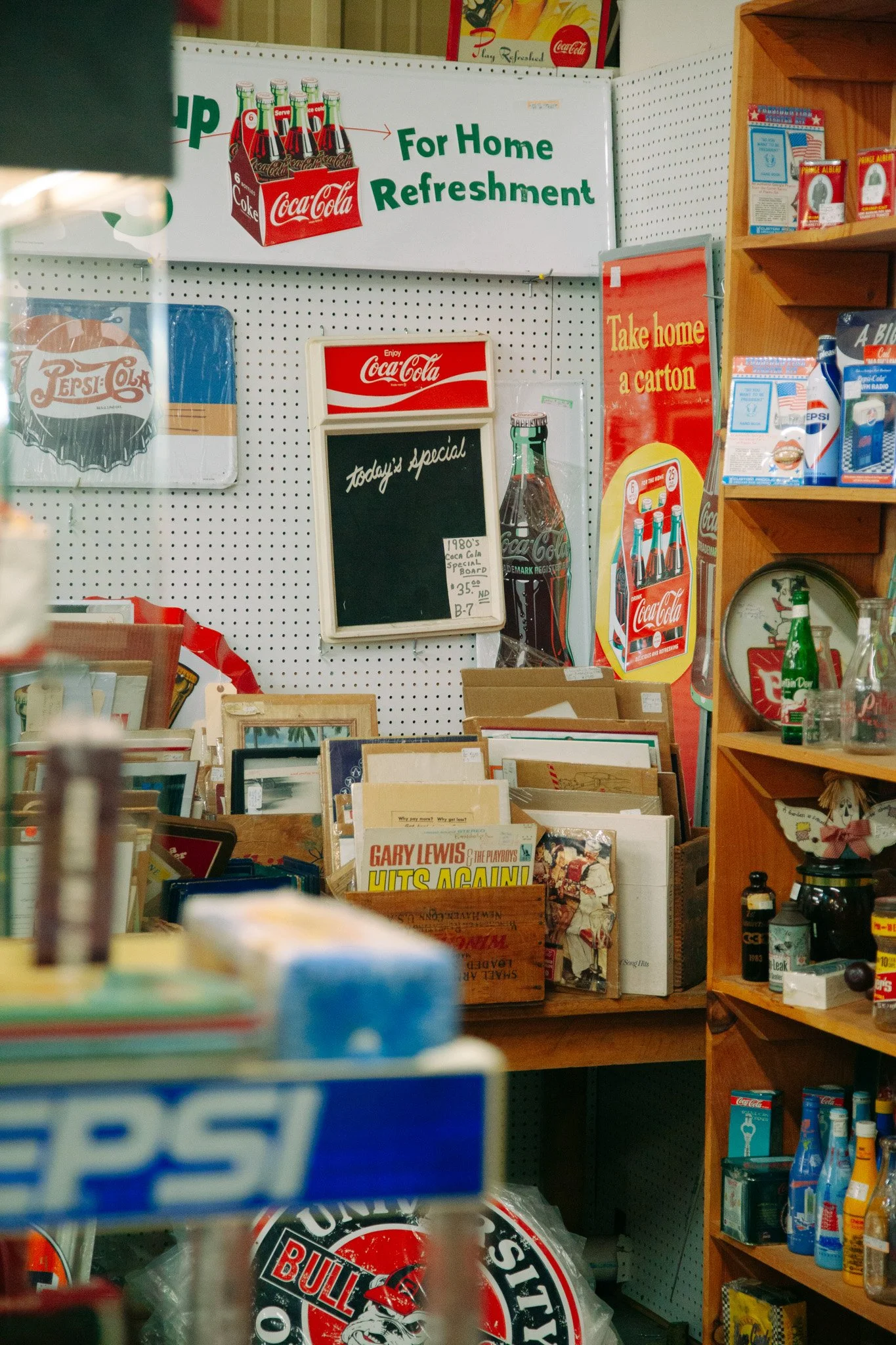 A retail store with vintage Coca-Cola signs, a chalkboard labeled 'Today's Special', and shelves filled with books and beverages. The main sign promotes Coca-Cola for home refreshment.
