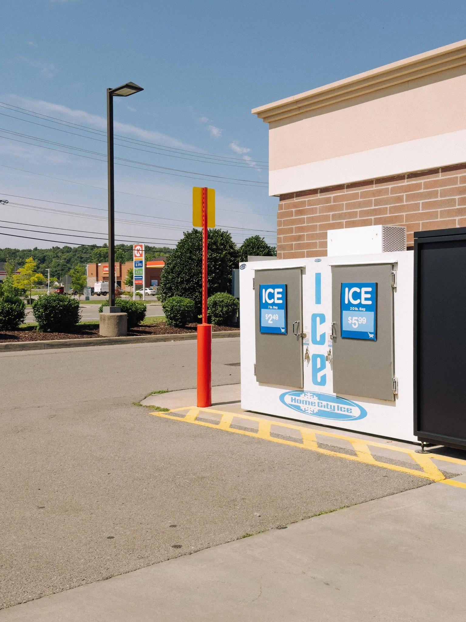 Empty outdoor ice vending machine with a blue and white logo, parking lot, trees, and shopping center in the background on a sunny day.