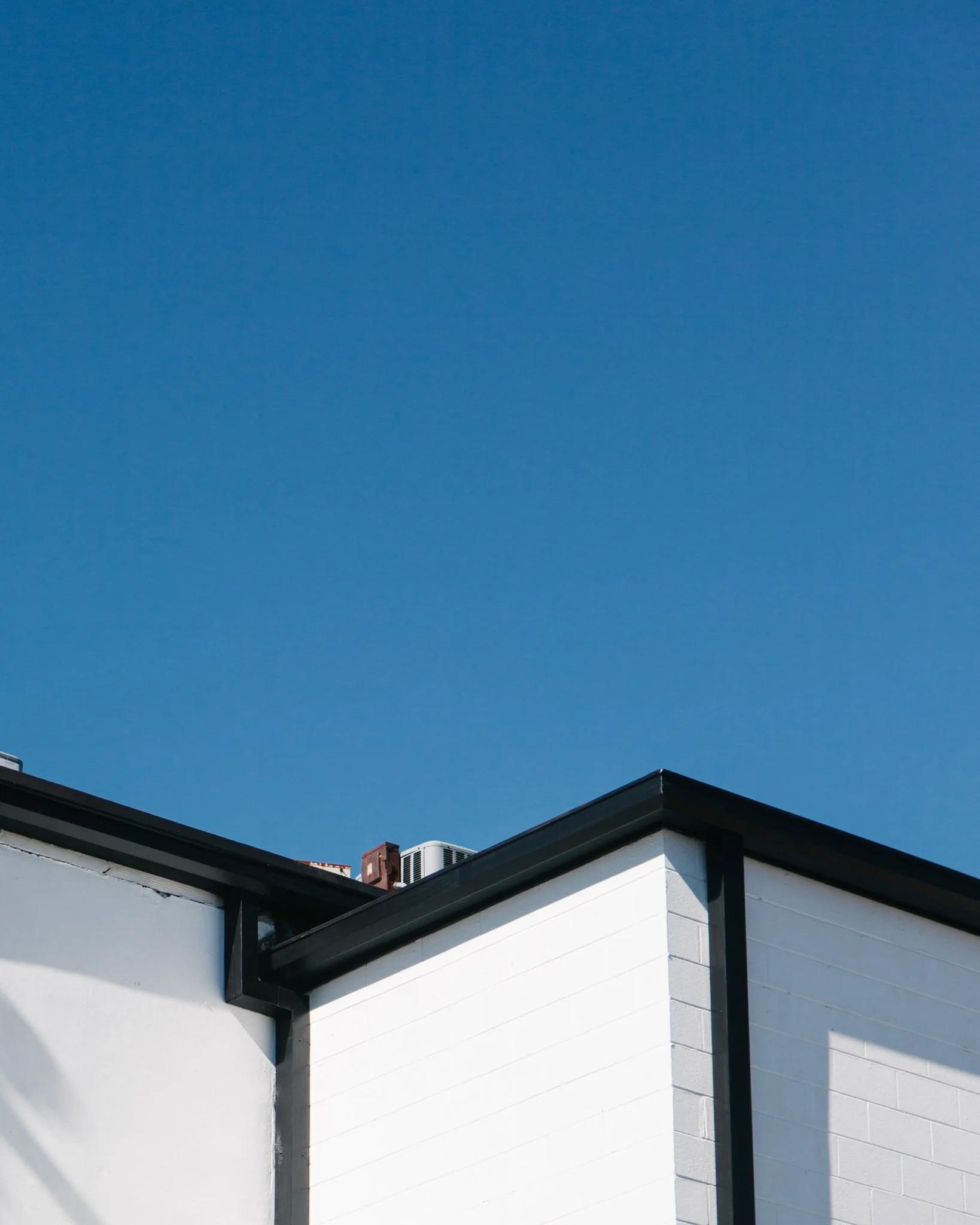 Close-up of the corner of a house with white painted walls, black trim, and a roof with dark shingles against a clear blue sky.