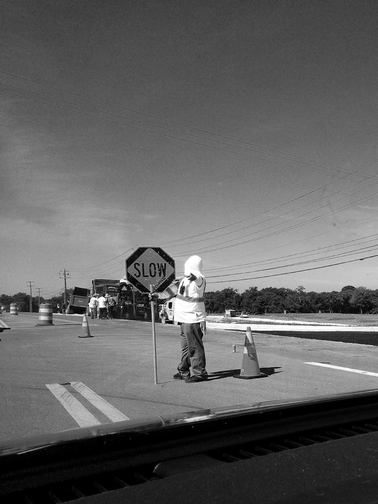 Workers in safety gear working on road construction, with traffic cones, a stop sign, and construction equipment on an open highway. The sky is clear with some power lines visible.
