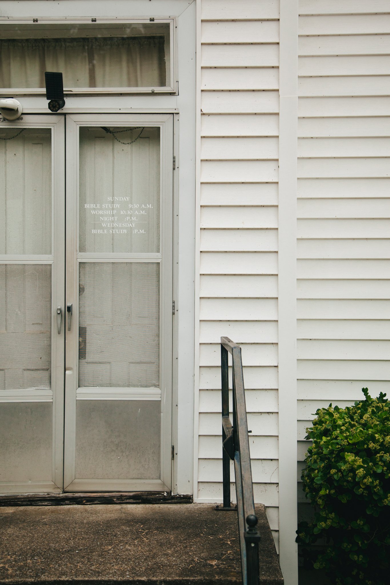 White exterior door with glass panels, a stair railing, and a bush on the right side, with white siding and a window above the door.