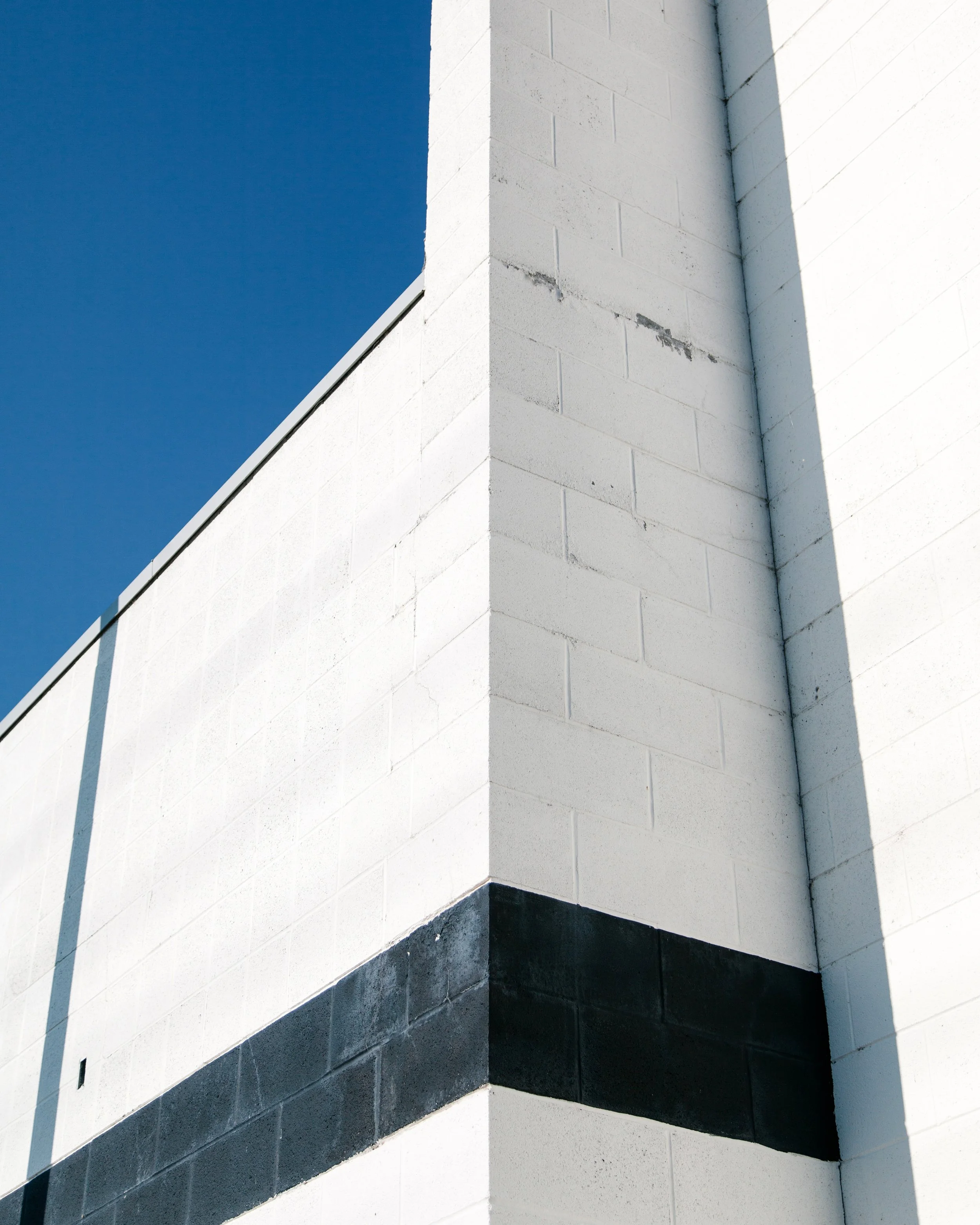 Close-up of a modern white building corner with a dark bottom section, under a clear blue sky, with shadows and slight weathering on the wall.