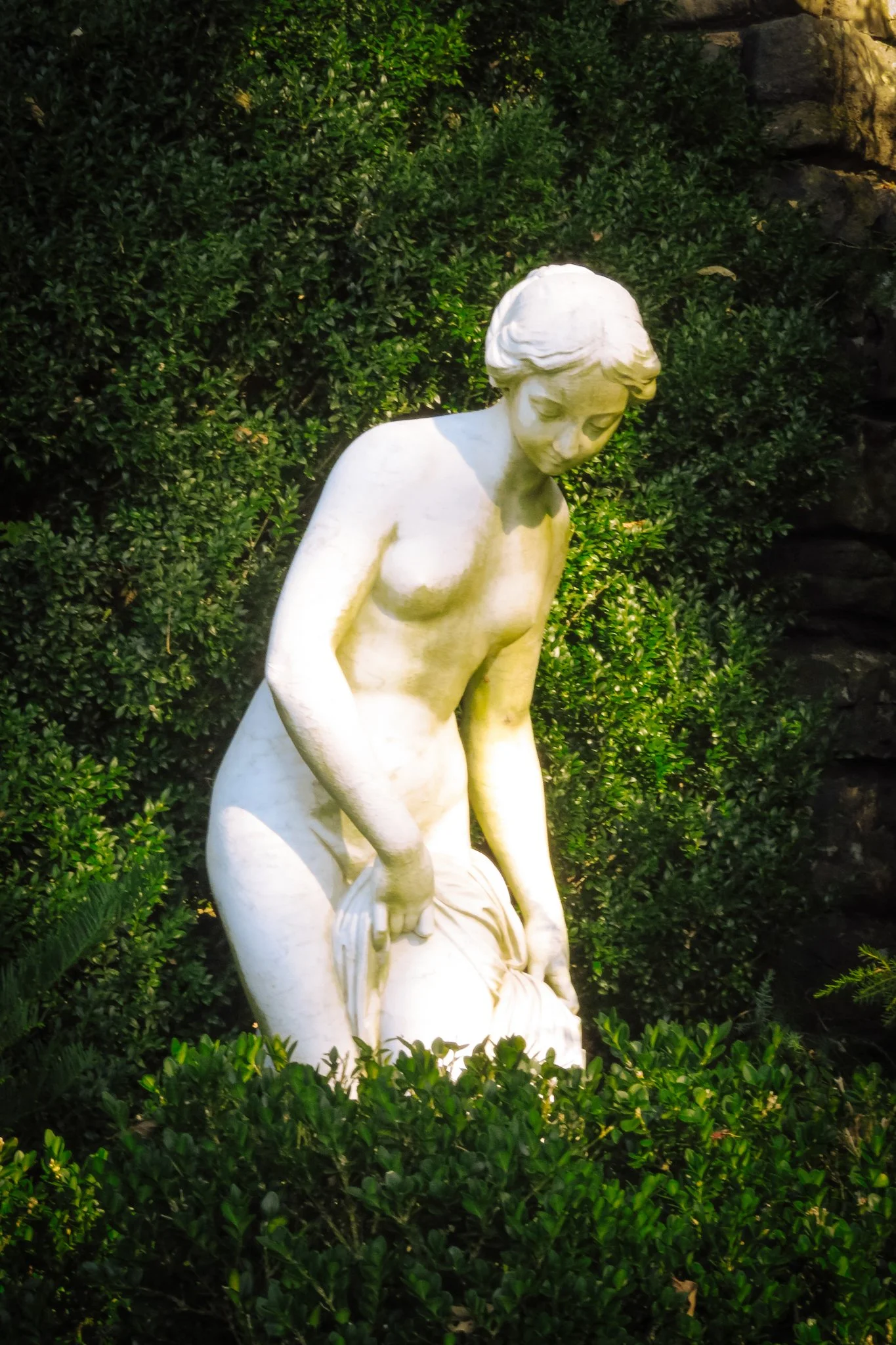 White marble statue of a woman in a garden with green bushes and rocks.
