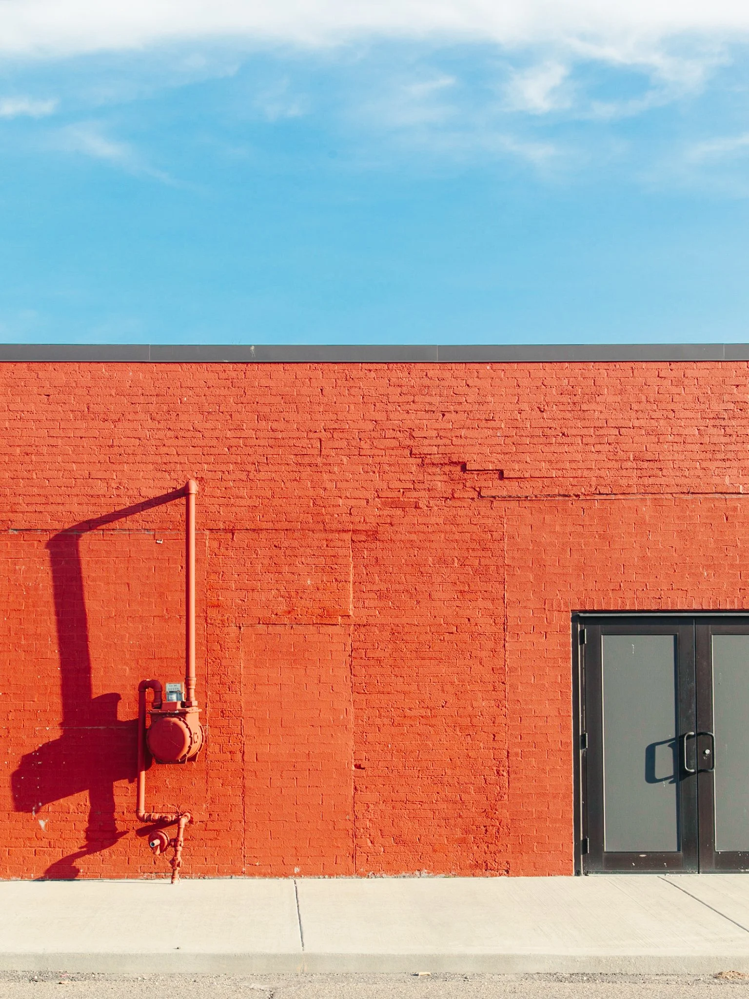 A red brick building wall with a black door on the right and a fire hydrant and pipes on the left, under a blue sky with some clouds.