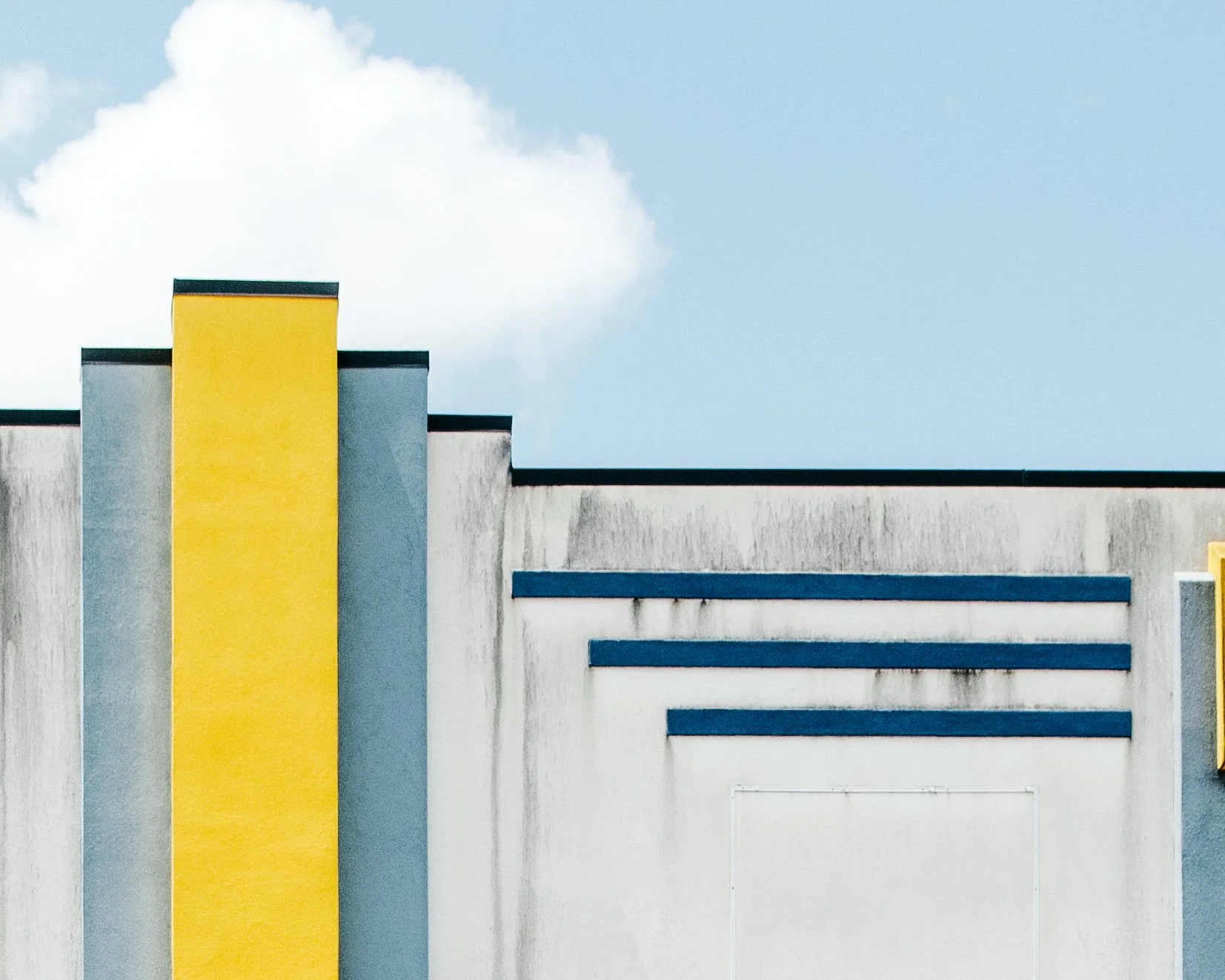Close-up of a modern building facade with yellow, gray, and blue elements under a partly cloudy sky.