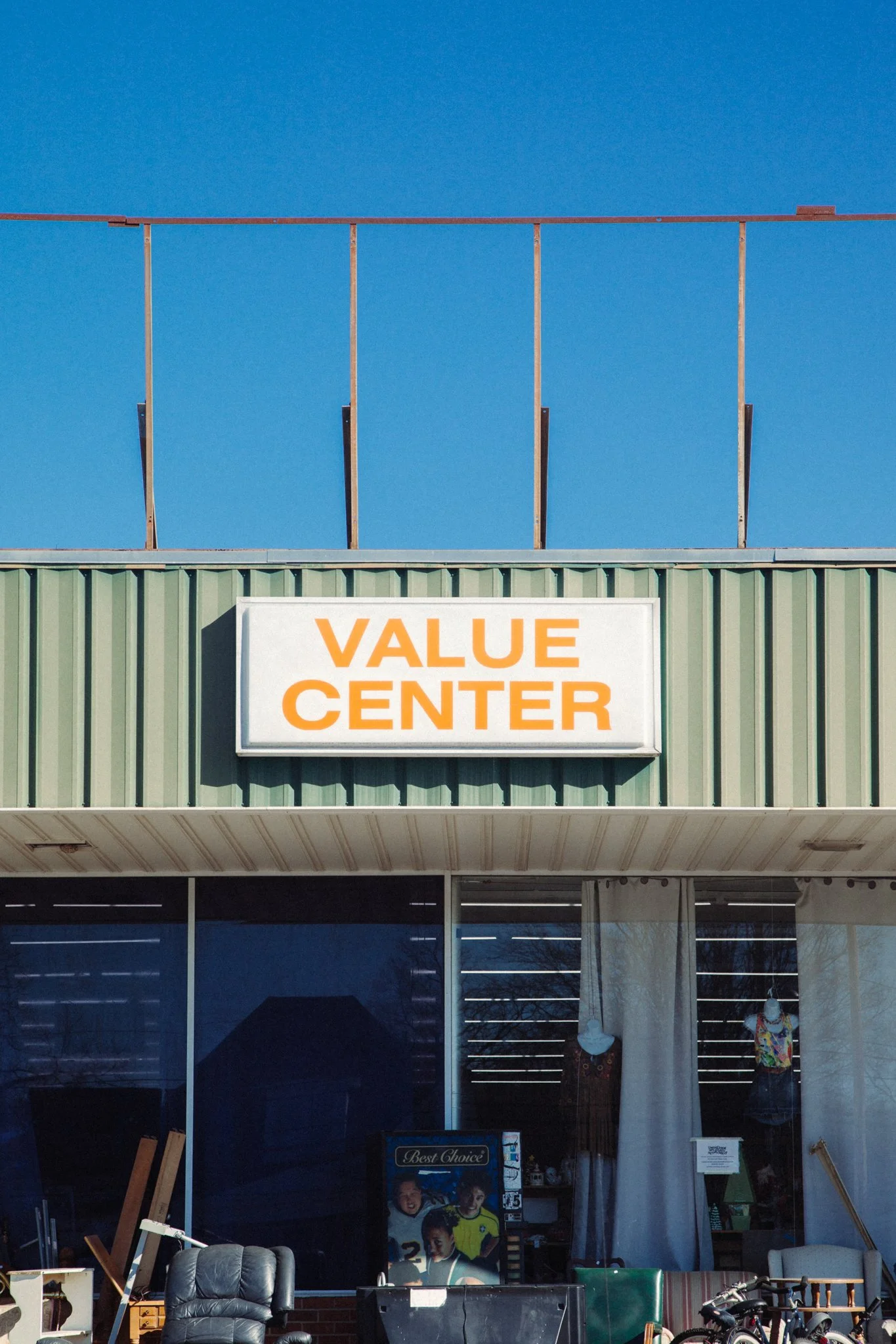 The exterior of a value center retail store with a large sign that reads 'VALUE CENTER'. The store has a green corrugated metal facade and large glass windows displaying mannequins dressed in various clothes. There are chairs, bicycles, and miscellan