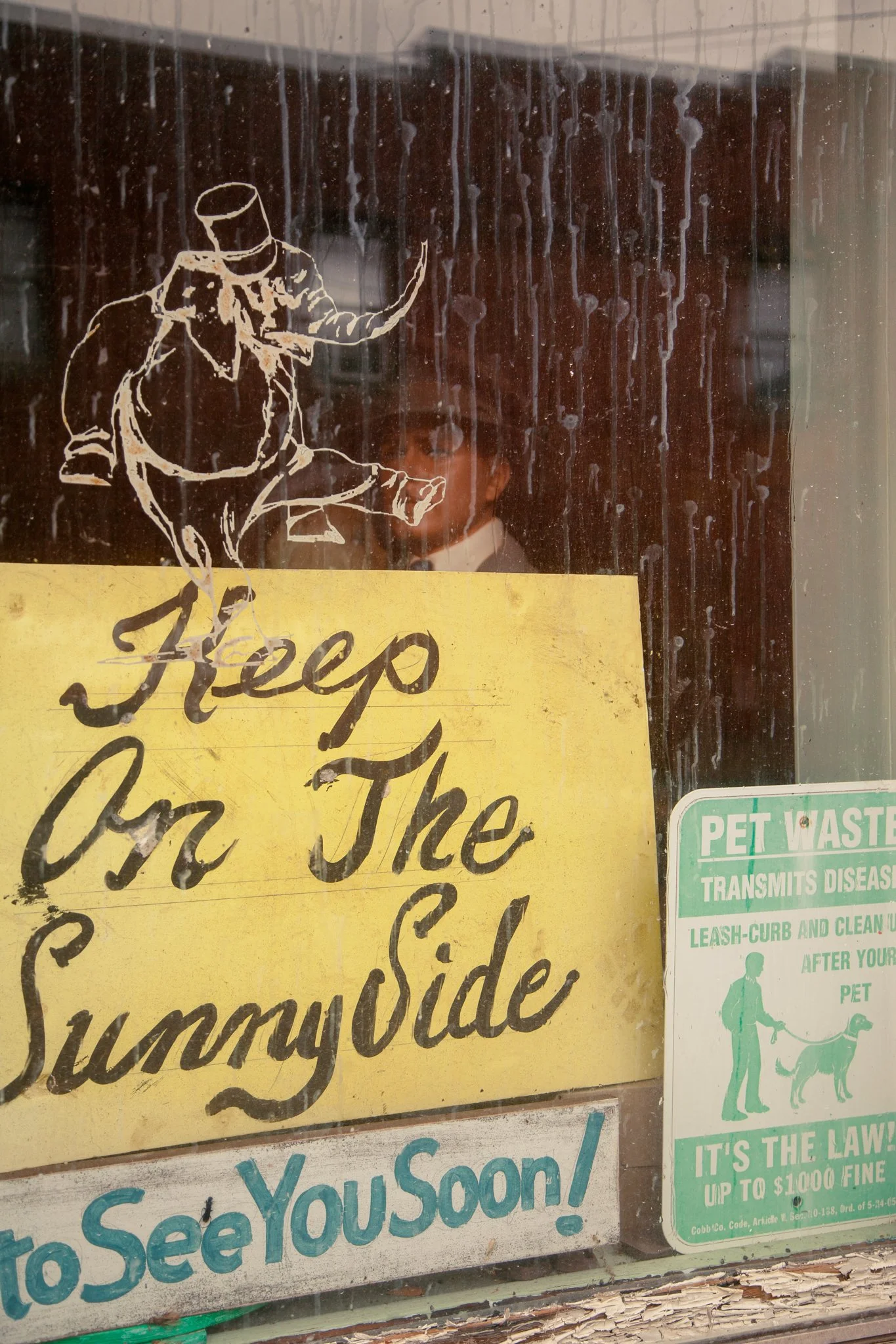 A glass window with a white outline of a man in a tuxedo and top hat, sitting with his legs crossed and waving, with a yellow sign below that says, 'Thank you for the sunshine.' There is a green pet waste disposal sign on the right side.