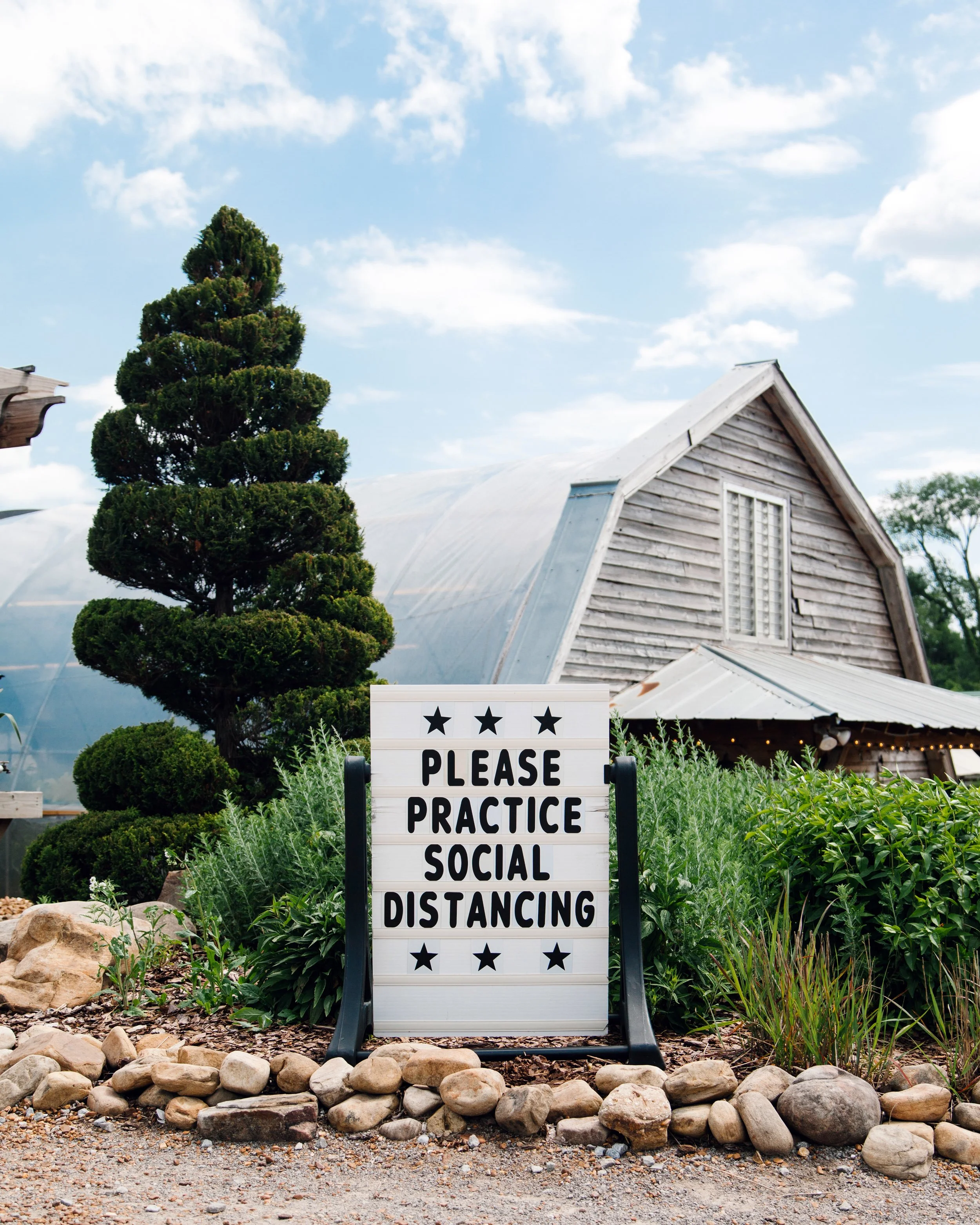 A sign with black letters on a white background displaying the message "Please Practice Social Distancing," surrounded by lush green plants and rocks, with a rustic building and a blue sky with clouds in the background.