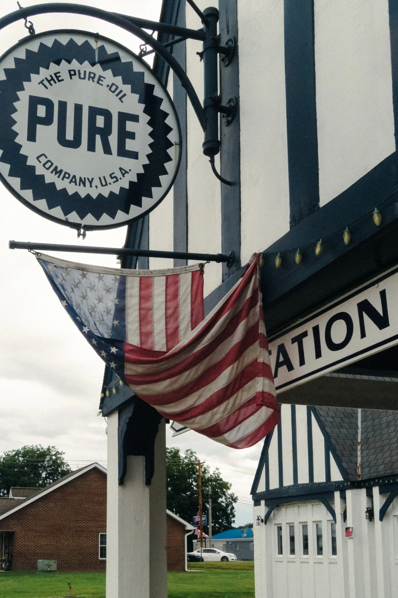 Store sign with the words 'Pure' and 'The Pure Oil Company, U.S.A.' hanging from the side of a building, with an American flag decoration below it and a white building with windows behind.