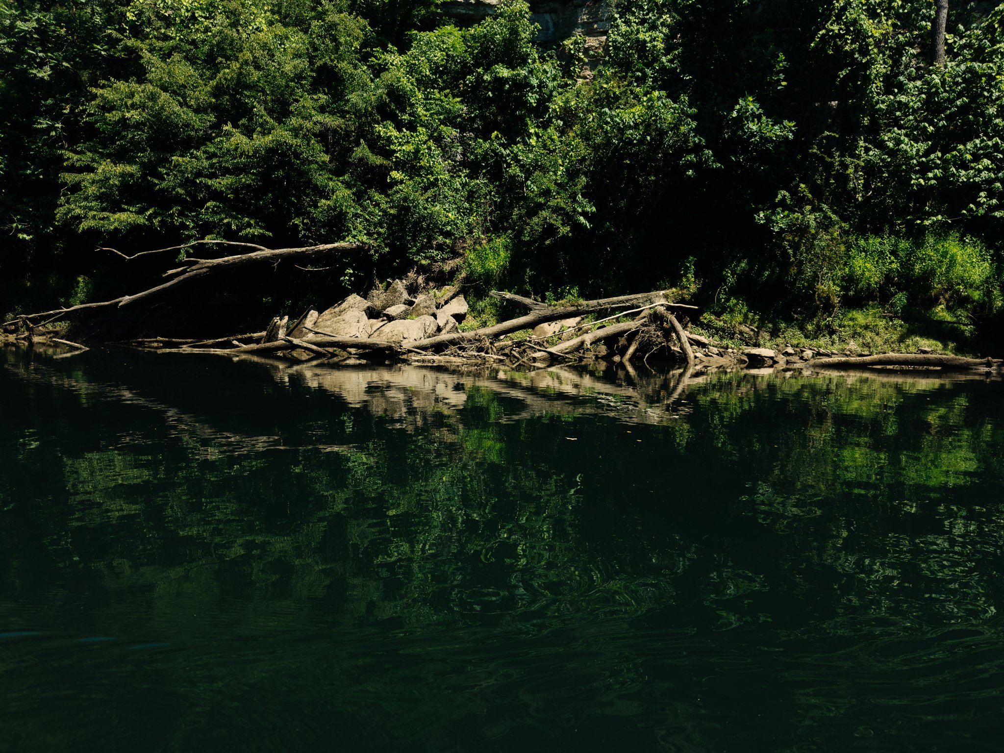 A lake with dark water, surrounded by rocks and dense green trees, reflecting in the water on a bright day.