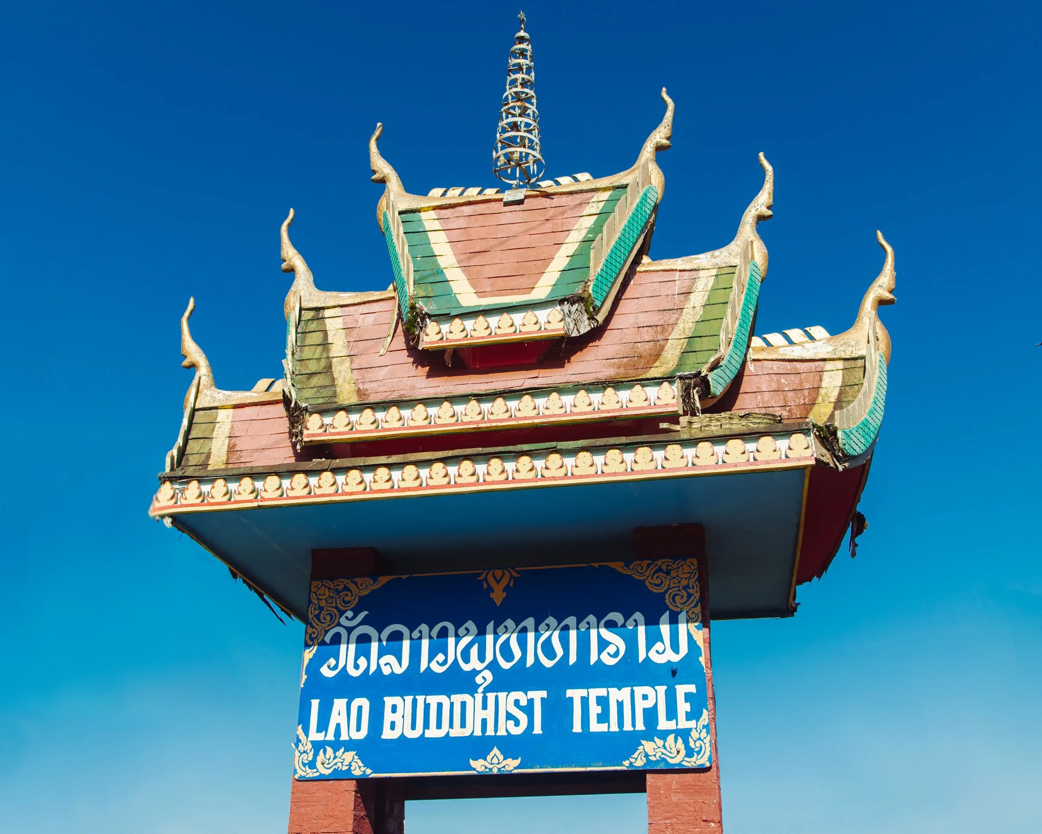Close-up of a colorful Lao Buddhist temple sign with traditional architectural details, against a clear blue sky.