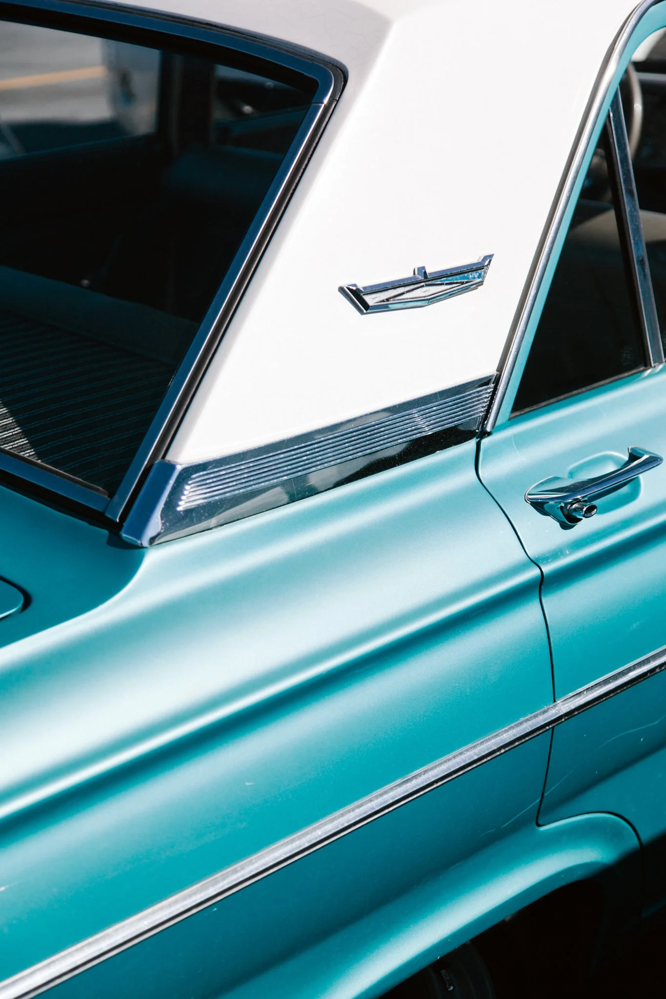 Close-up of a vintage light blue and white car, showing the side window, door handle, and a chrome emblem on the side.