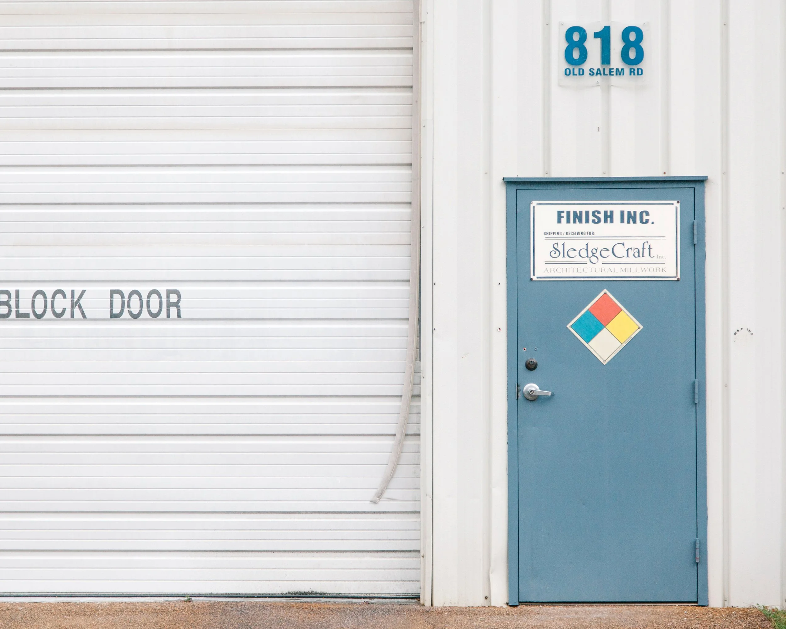 White industrial building with a large closed white roll-up door and a blue door next to it. The blue door has a sign that reads "Finish Inc." The building has the address "818 Old Salem Rd" displayed in blue numbers and letters on the wall near the 
