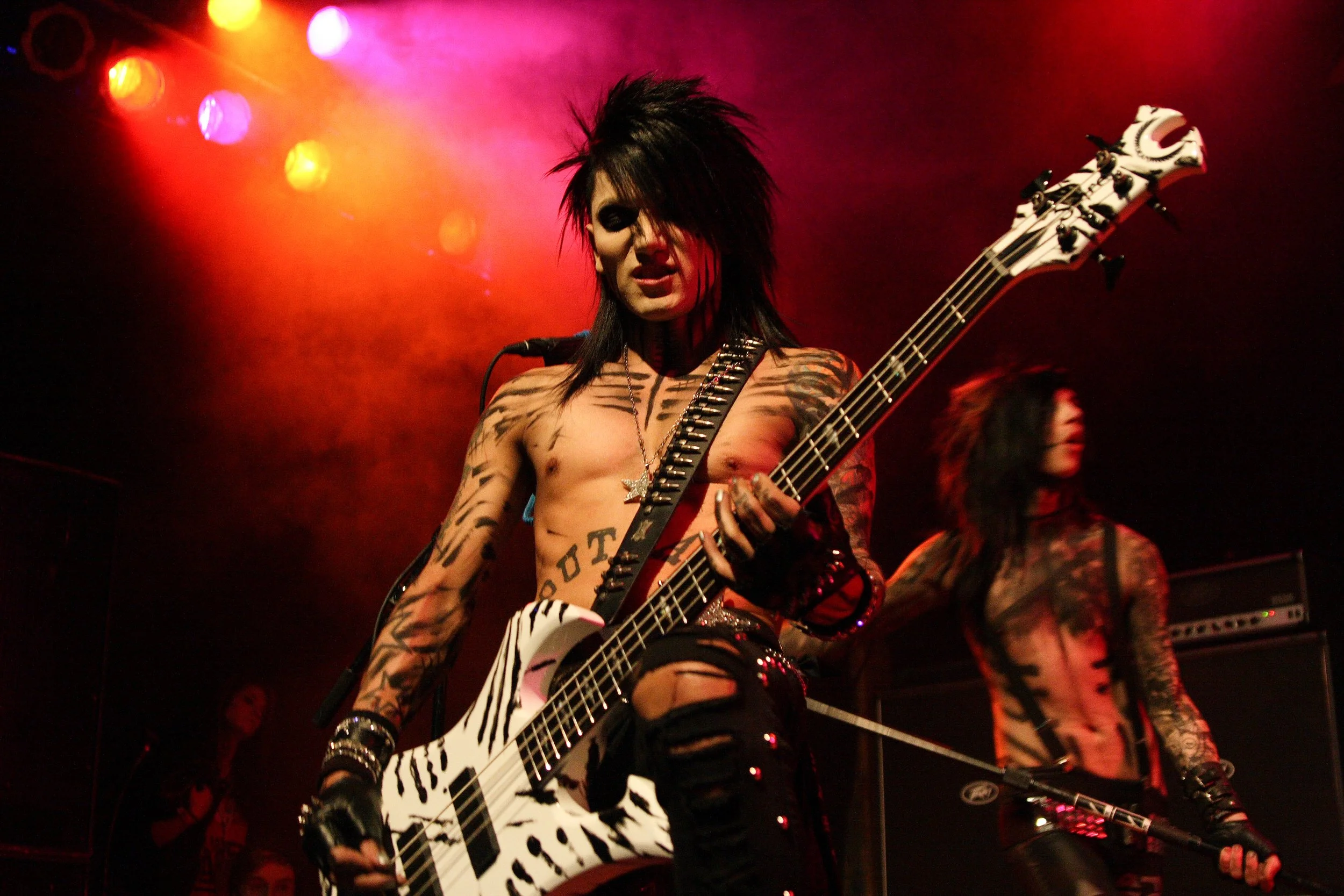 A shirtless punk rock musician with tattoos and spiked black hair playing an electric guitar on stage with colorful lights in the background.