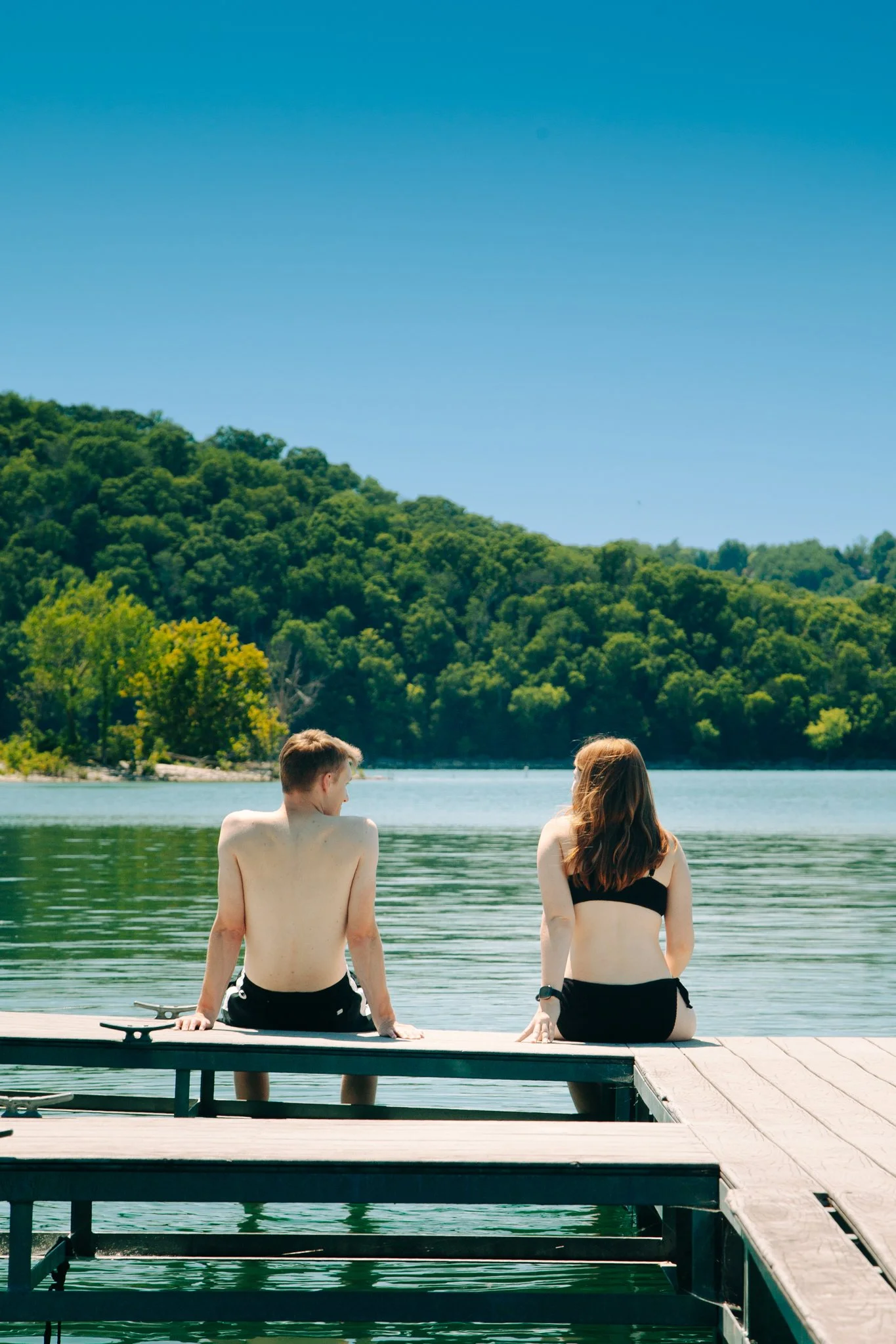 A young man and woman sit on a dock at a lake, facing the water with their backs to the camera, with lush green trees and a clear blue sky in the background.