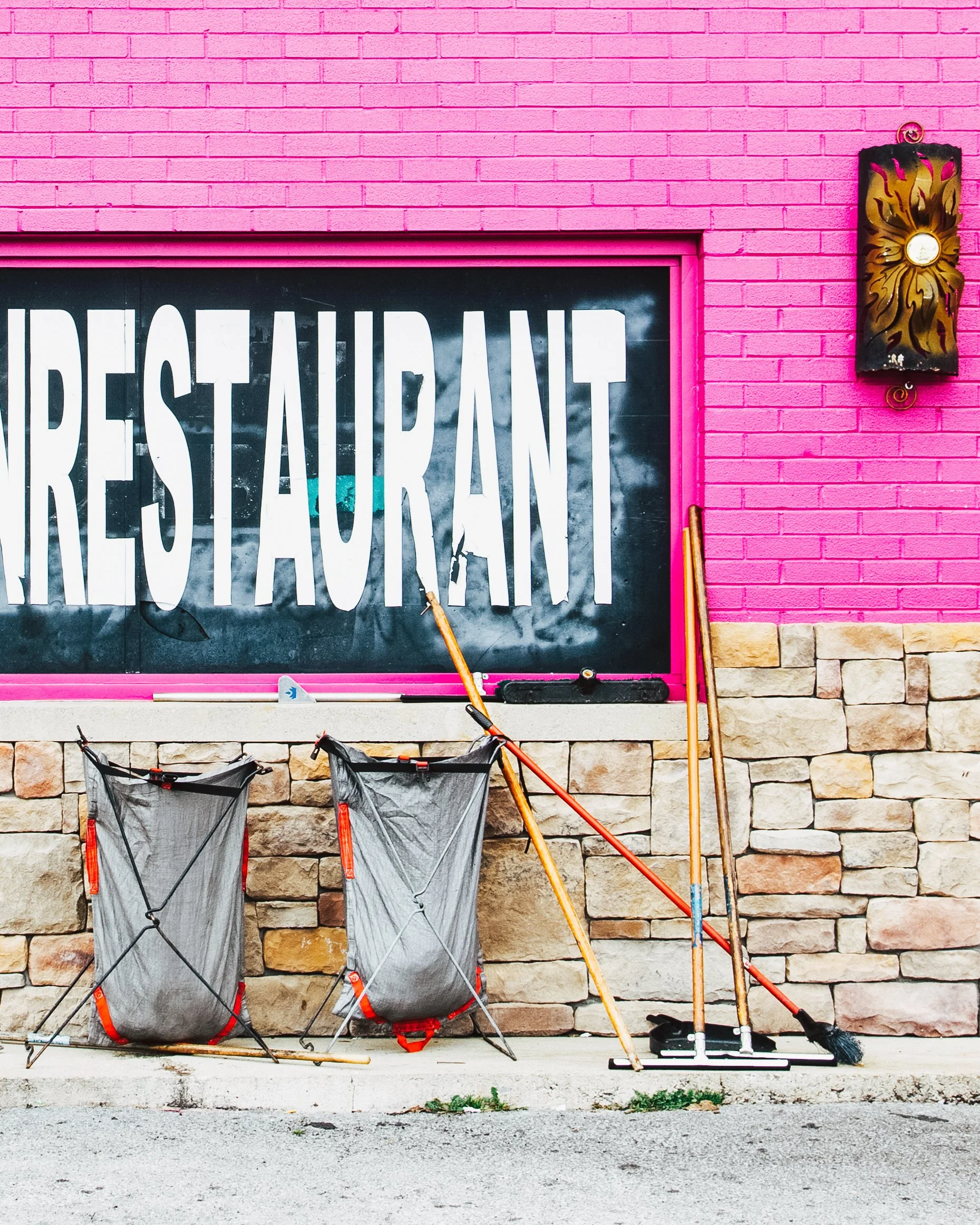 A pink brick wall with a large window displaying the word 'RESTAURANT' and cleaning equipment including two laundry baskets, brooms, and a mop placed outside along the street.